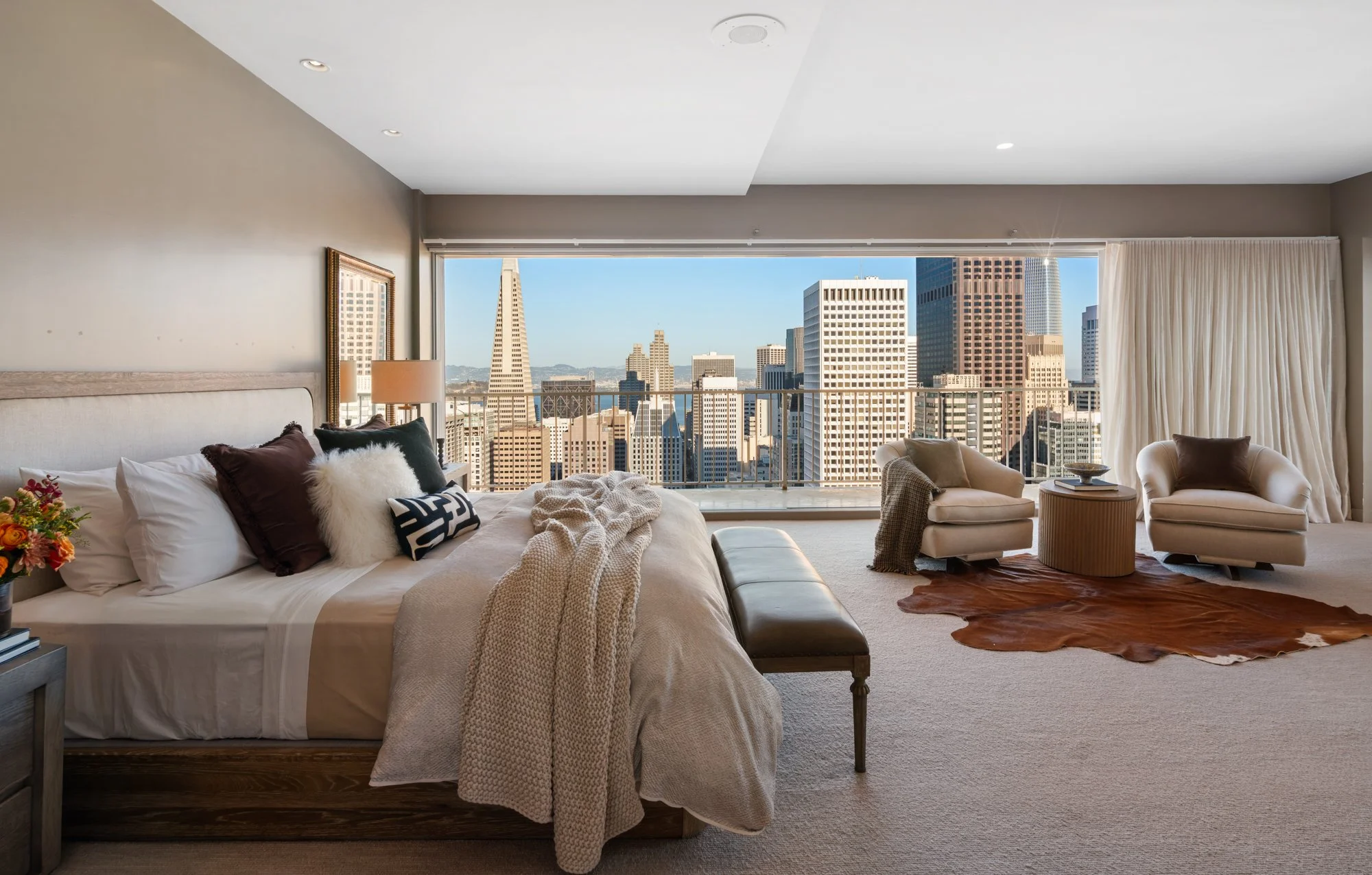A spacious modern living room with large windows overlooking San Francisco's skyline, including the Transamerica Pyramid. The room features a plush bed with pillows, a cozy throw blanket, a small side table with a flower arrangement, and a seating ar