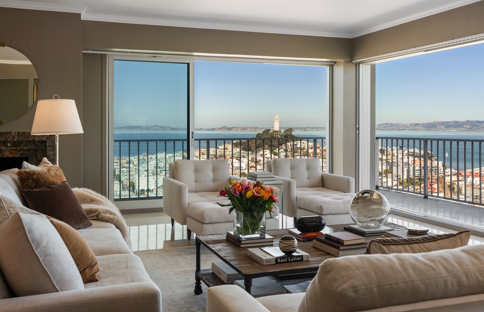 Living room with large windows revealing a city and water view, furnished with white sofas, a coffee table with books and decorative items, and a choice of pillows and throws.