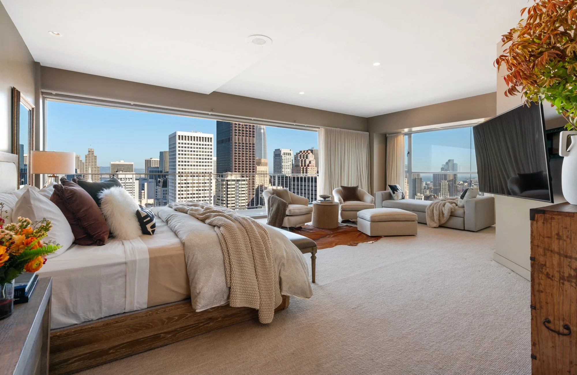 A spacious living room with large windows offering a city skyline view. The room features a bed with pillows and blankets, a wooden side table, several armchairs, a flat-screen TV, and decorative plants.