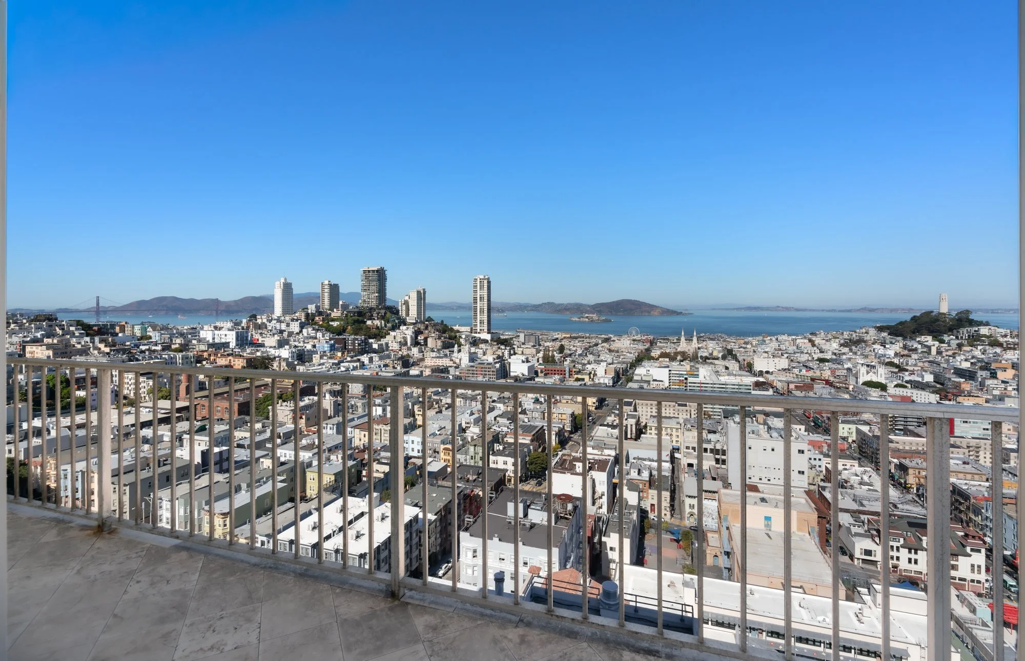 View from a balcony overlooking a cityscape with tall buildings, a water body, hills, and a bridge in the background under a clear blue sky.