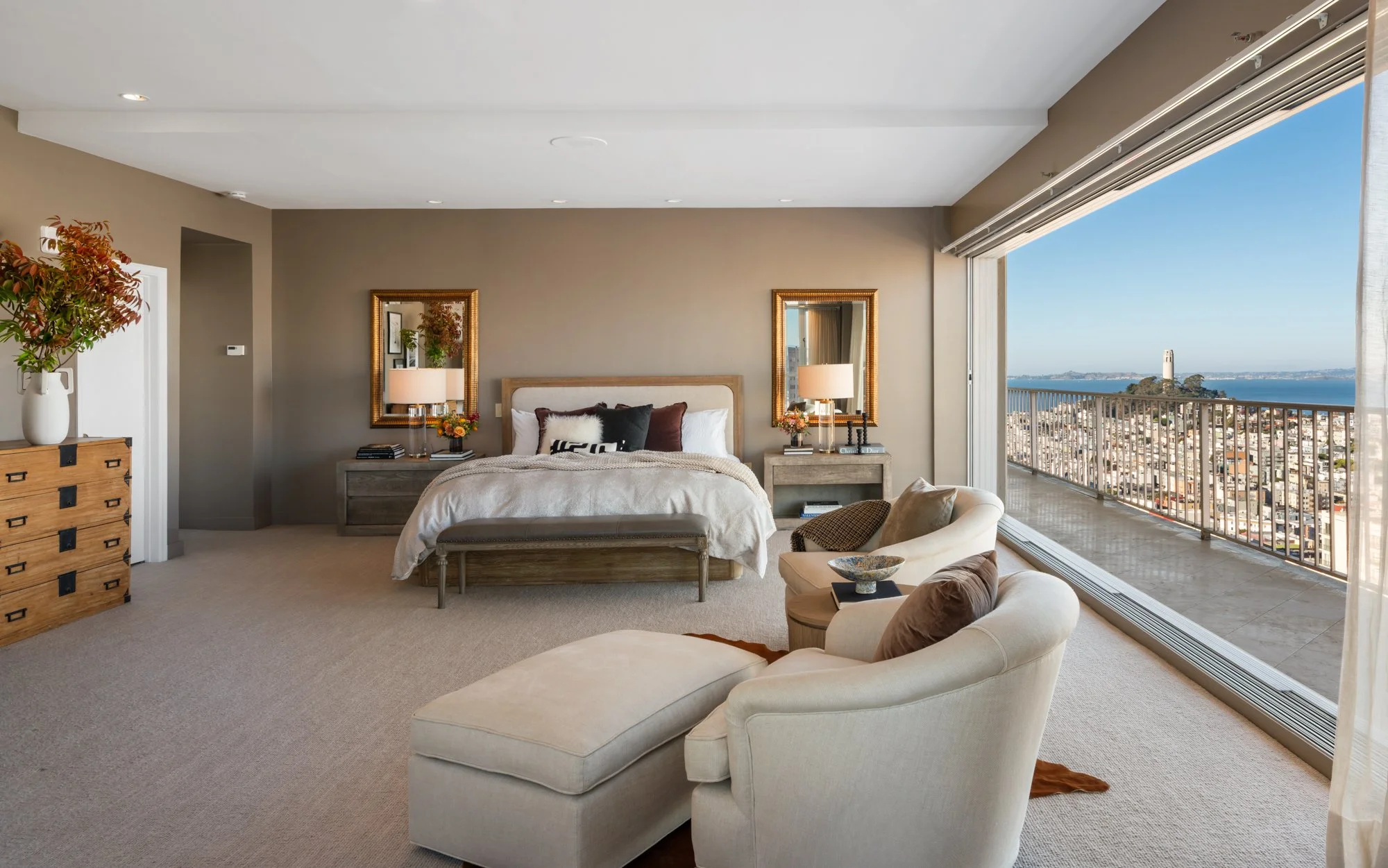 Modern bedroom with large window overlooking city and water, neutral-colored furniture including a bed, armchair, and dresser, decorated with lamps, mirrors, and plants.