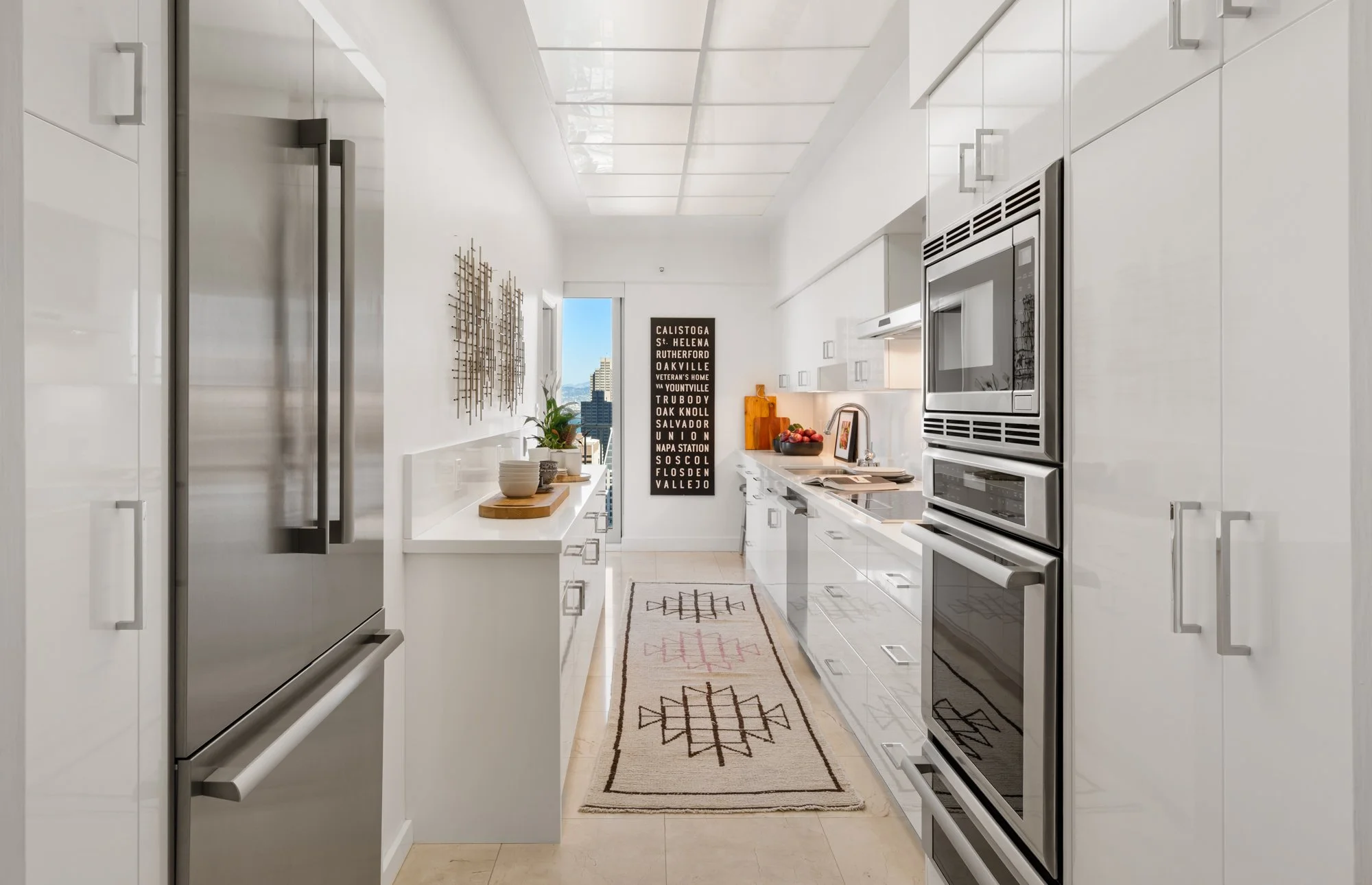 Modern white kitchen with stainless steel appliances, including a large refrigerator on the left and a double oven on the right, with a decorative wall hanging and multiple potted plants on a white countertop.
