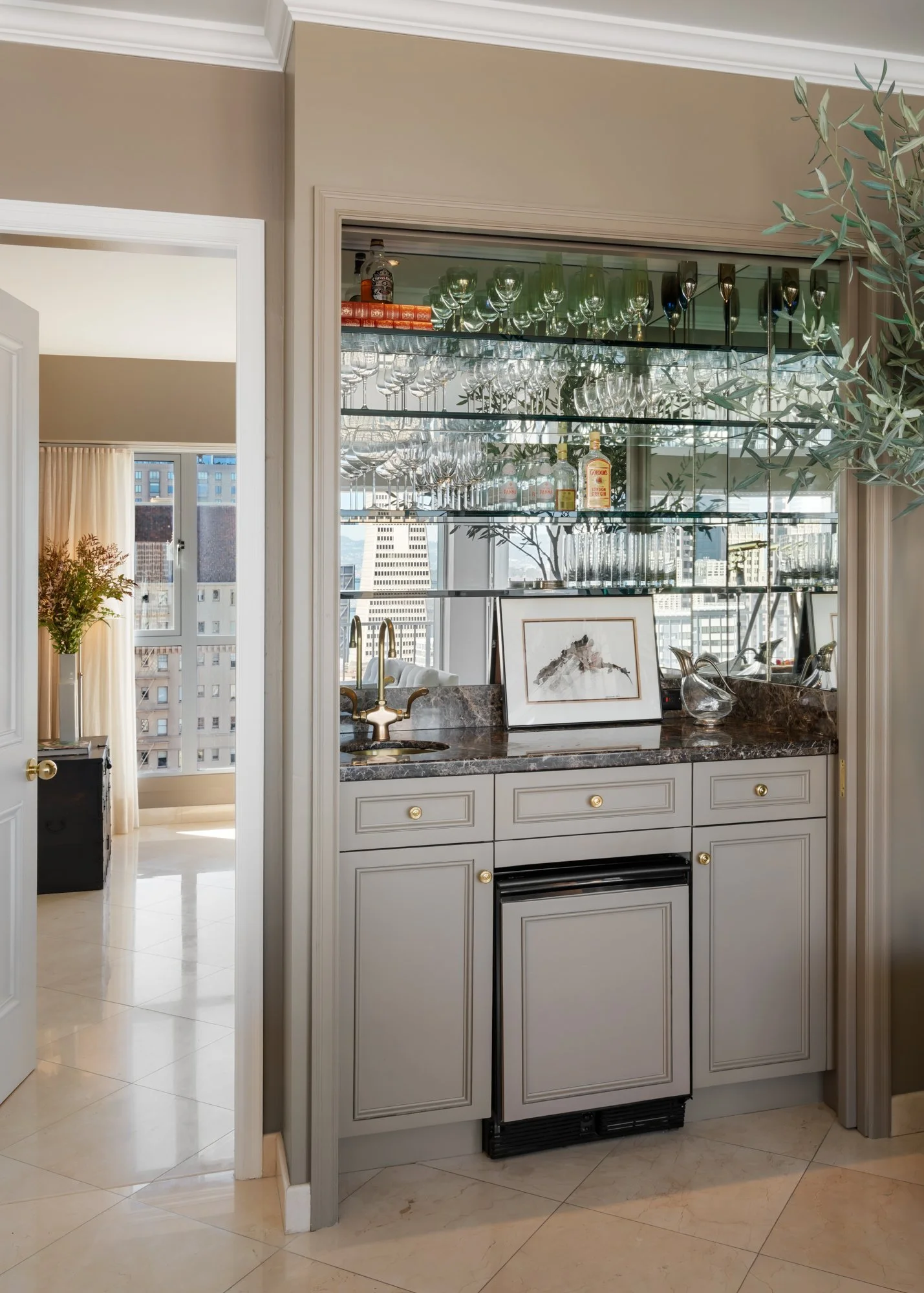 A modern bar area inside a high-rise apartment featuring a mirrored glass shelving unit with wine glasses and liquor bottles, a marble countertop with a framed artwork and decorative glass pitcher, and city skyline views through large windows.