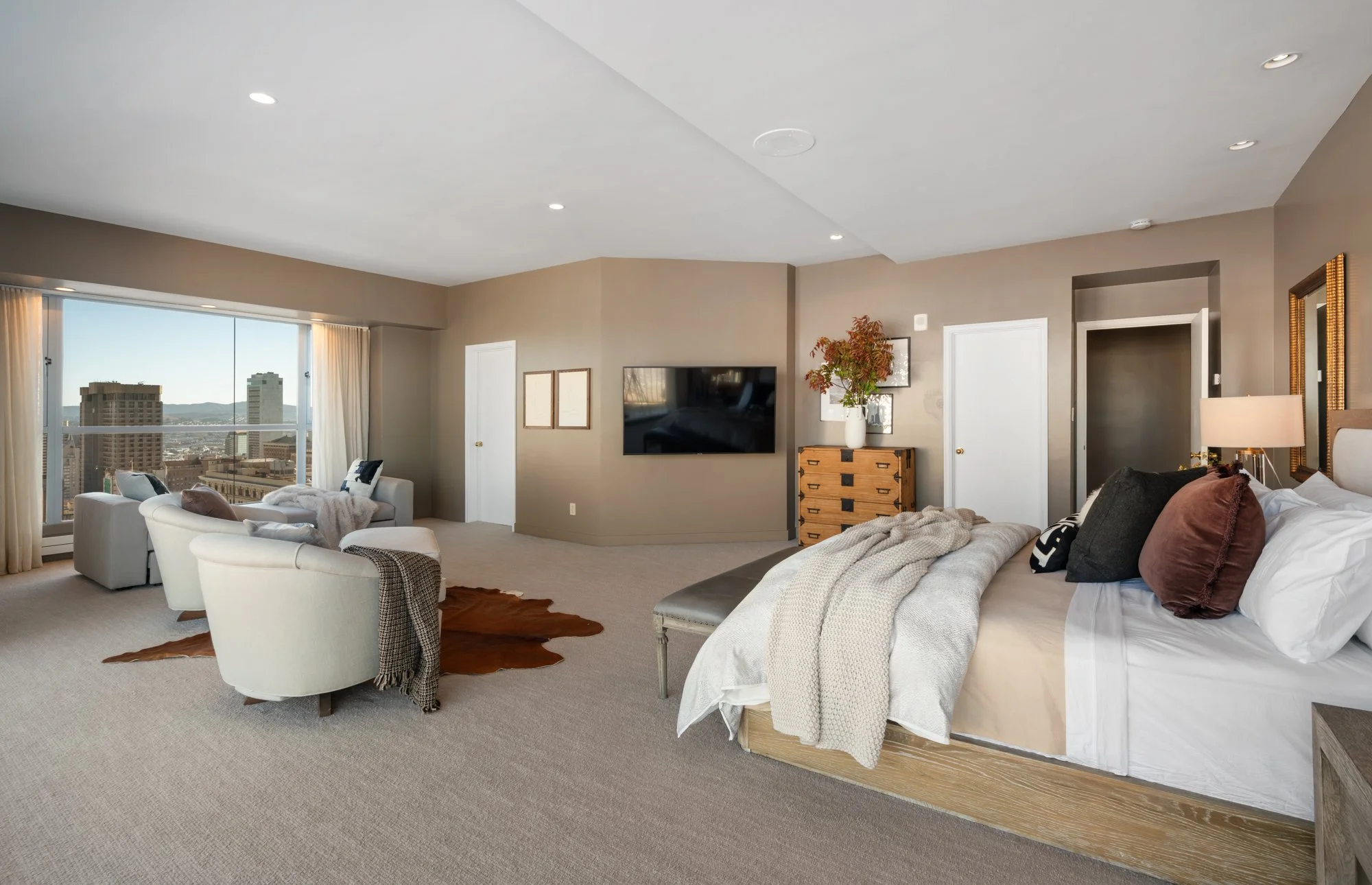 A spacious living room with beige walls and a large window showing city buildings. The room has white armchairs, a leather bench, a TV on the wall, and a bedroom area with a bed and pillows. There is a wooden dresser with a plant and framed pictures,