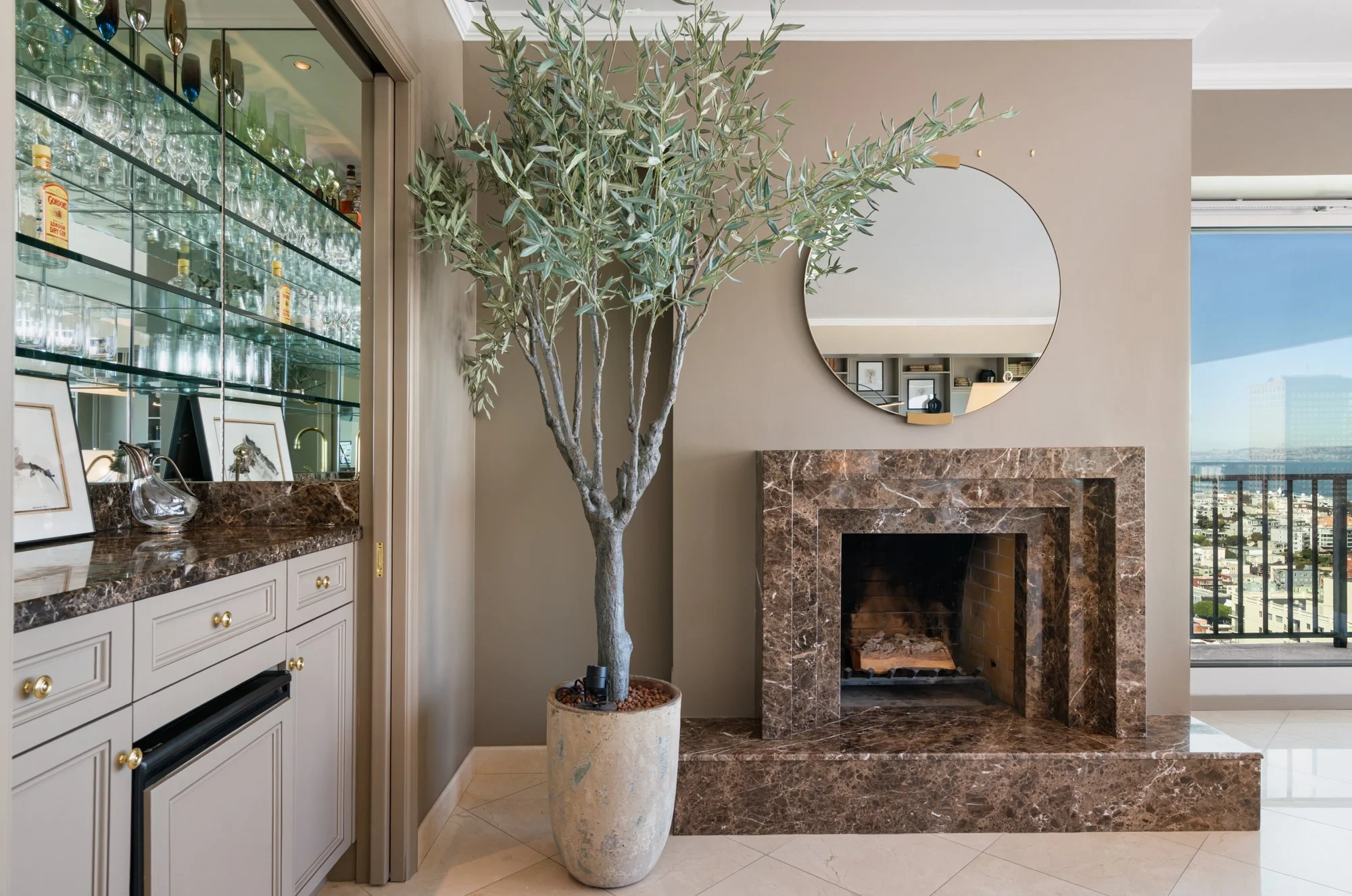 Indoor space with a large potted olive tree, white cabinetry, marble fireplace, and a round mirror on beige wall, with city skyline visible through a window.