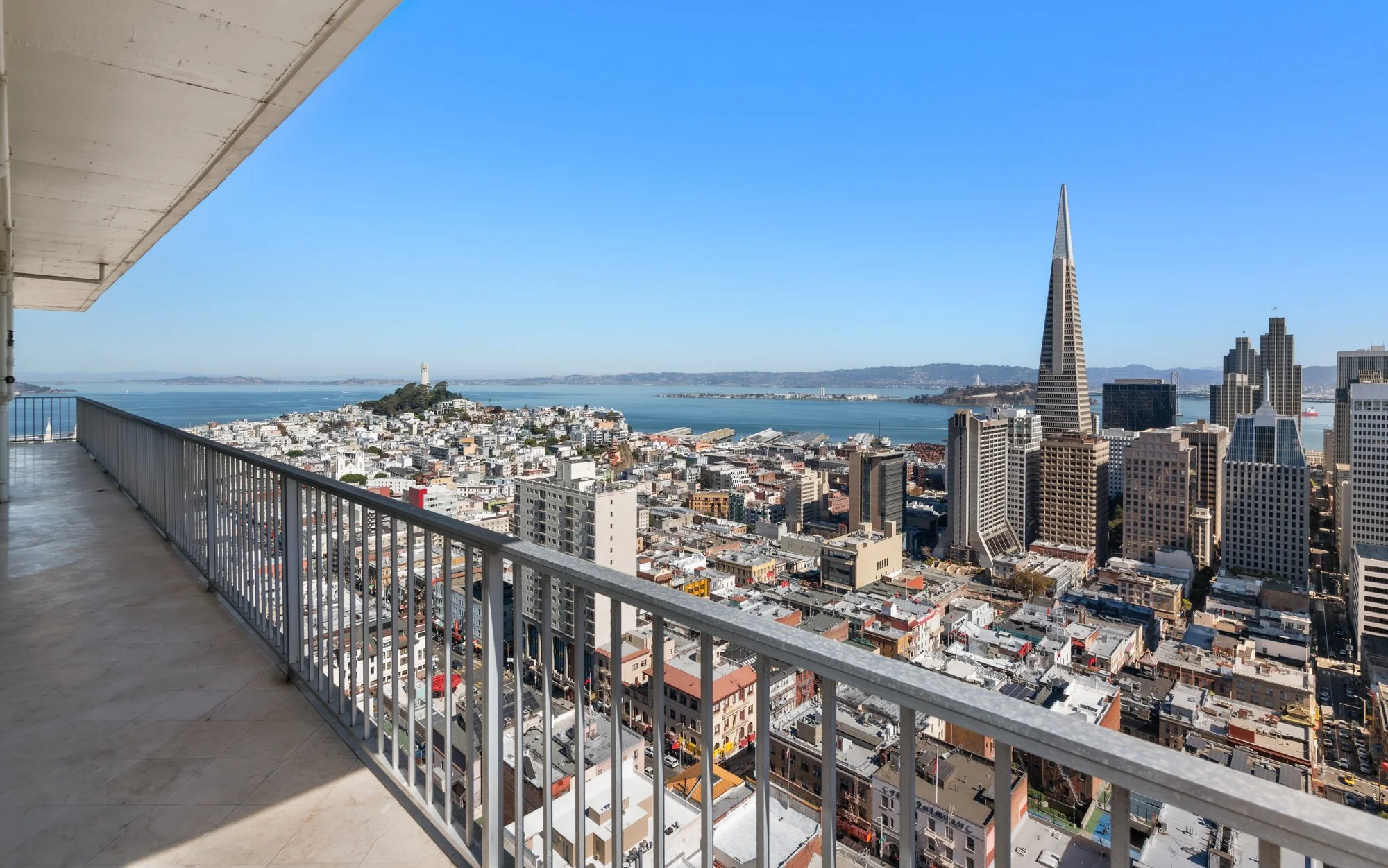 City skyline view from a high-rise balcony on a clear day, featuring the Transamerica Pyramid and San Francisco Bay, with a blue sky and distant landforms.