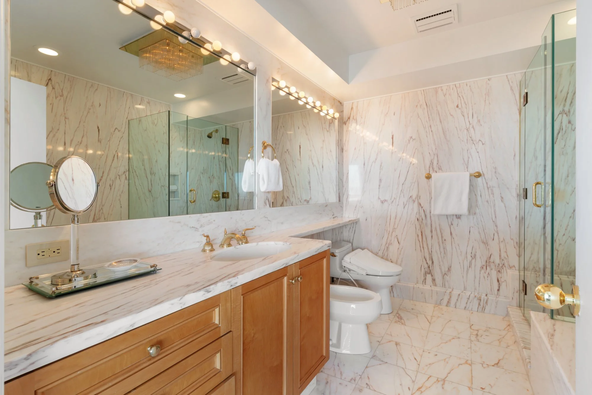 A luxurious bathroom with marble walls and floor, a large mirror with globe lights, a marble countertop with a sink and vanity, a toilet, and a glass shower enclosure with a gold handle.