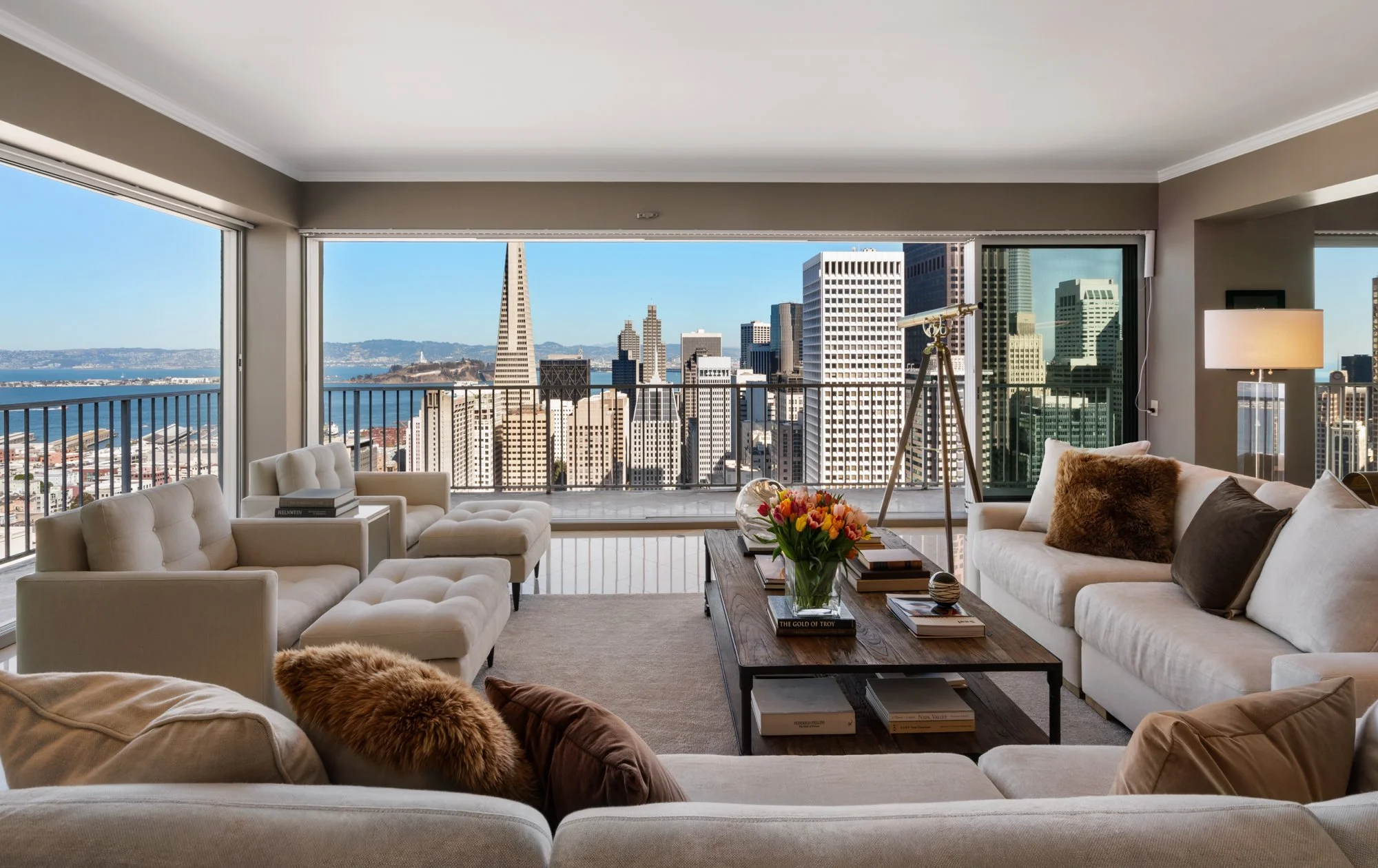 Living room with large windows showing cityscape with skyscrapers, including the Transamerica Pyramid, and water. The room has beige and white furniture, a wooden coffee table with books and a flower vase, and a telescope.