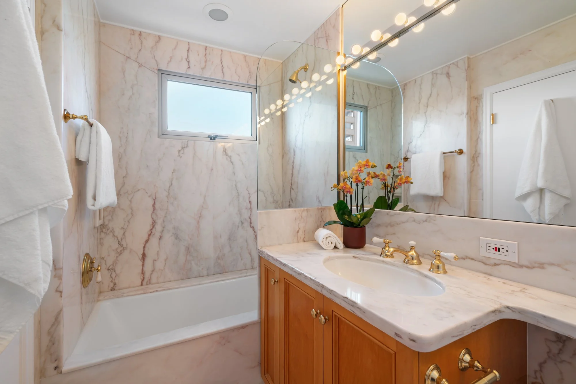 A bathroom with marble walls and countertop, a built-in bathtub, a wooden vanity with a marble top, a potted orchid on the counter, and towels hanging on gold fixtures. There is a window and a large mirror above the sink.
