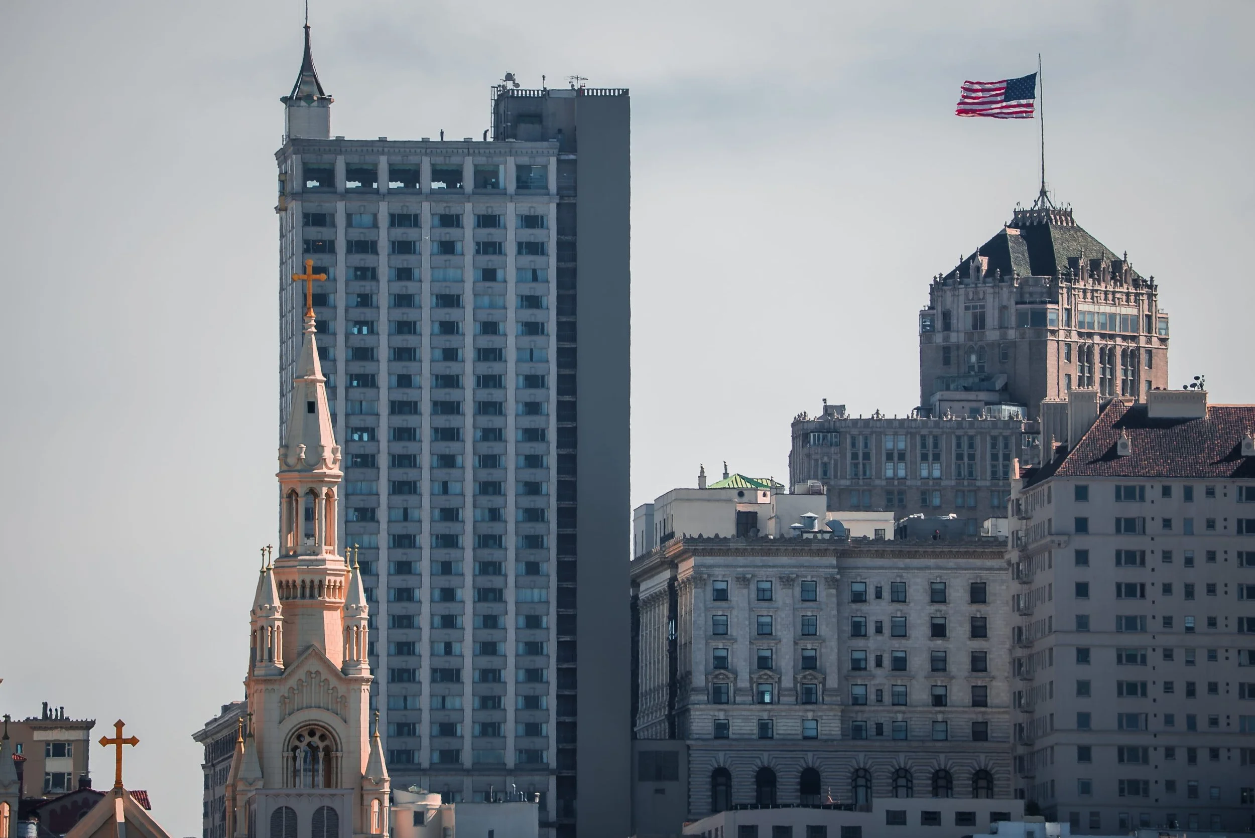 A city skyline with a church steeple in front of tall modern buildings, including a skyscraper with a small tower on top and an American flag flying on a rooftop.