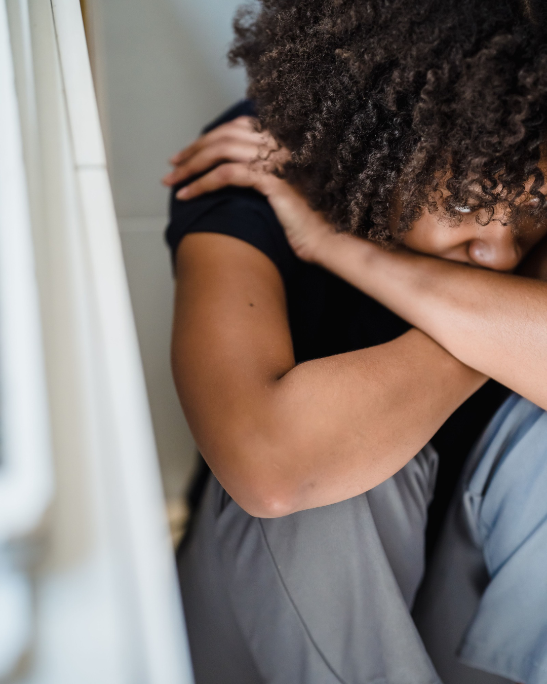 A person in emotional distress sitting with their head down, hugging their knees, and holding their shoulder with one hand.