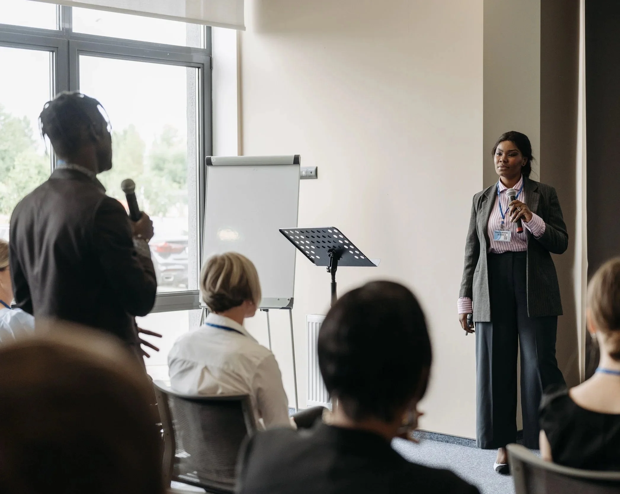 A woman in a gray suit and pink shirt holding a microphone stands in front of a audience, speaking during a presentation or conference in a bright room with large windows.