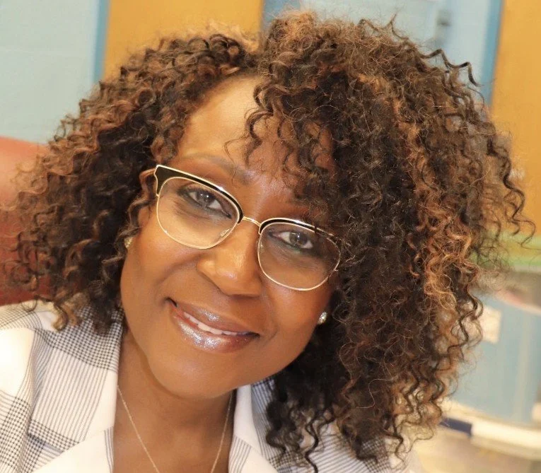 A woman with curly hair wearing glasses and a striped blazer, smiling at the camera.