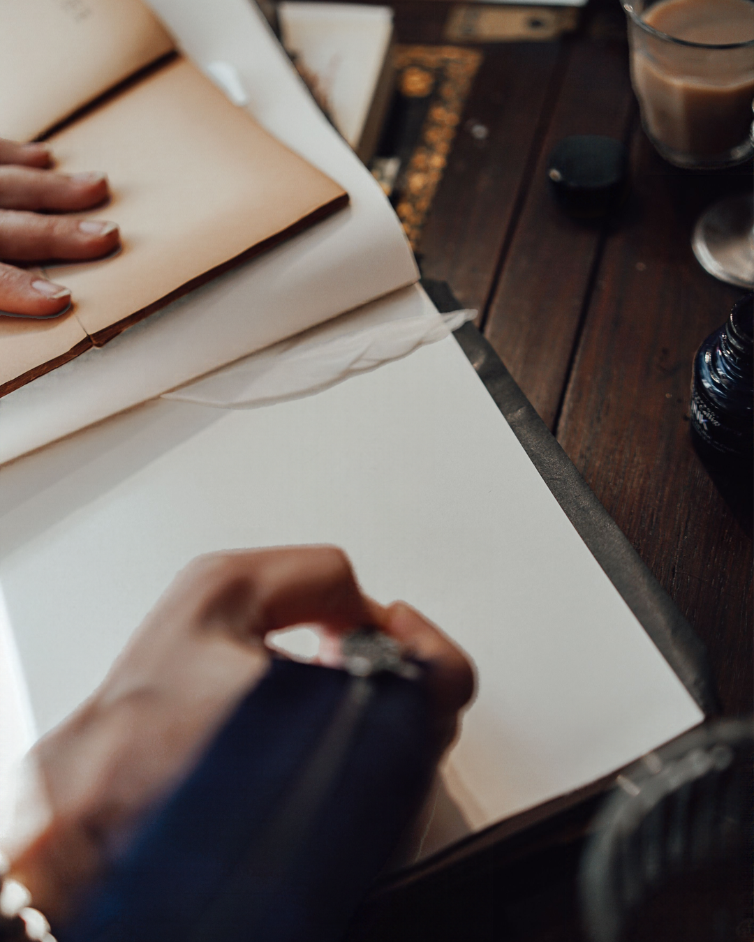 A person writing with a pen on a blank sheet of paper on a wooden table, with a book and drink nearby.