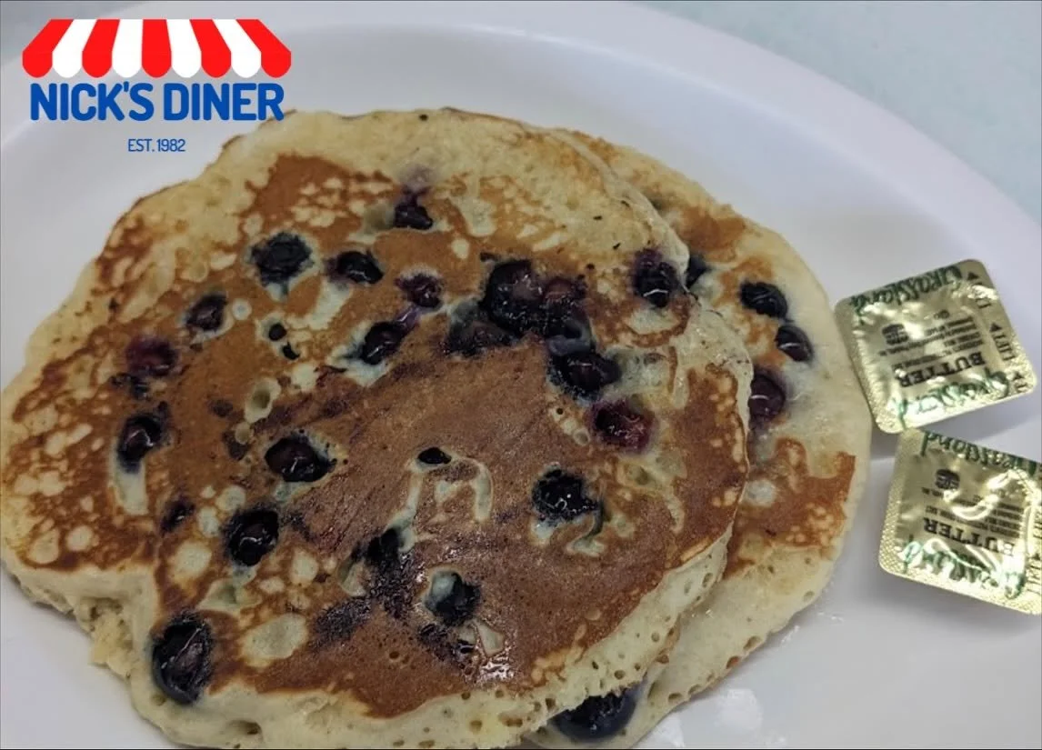 Two blueberry pancakes topped with cinnamon on a white plate, with two packets of butter on the side, branded Nick's Diner established 1982.