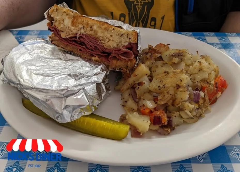 A plate of food with a partially unwrapped sandwich with roast beef and bread, a scoop of potato salad with vegetables, a dill pickle spear, and in a restaurant setting.