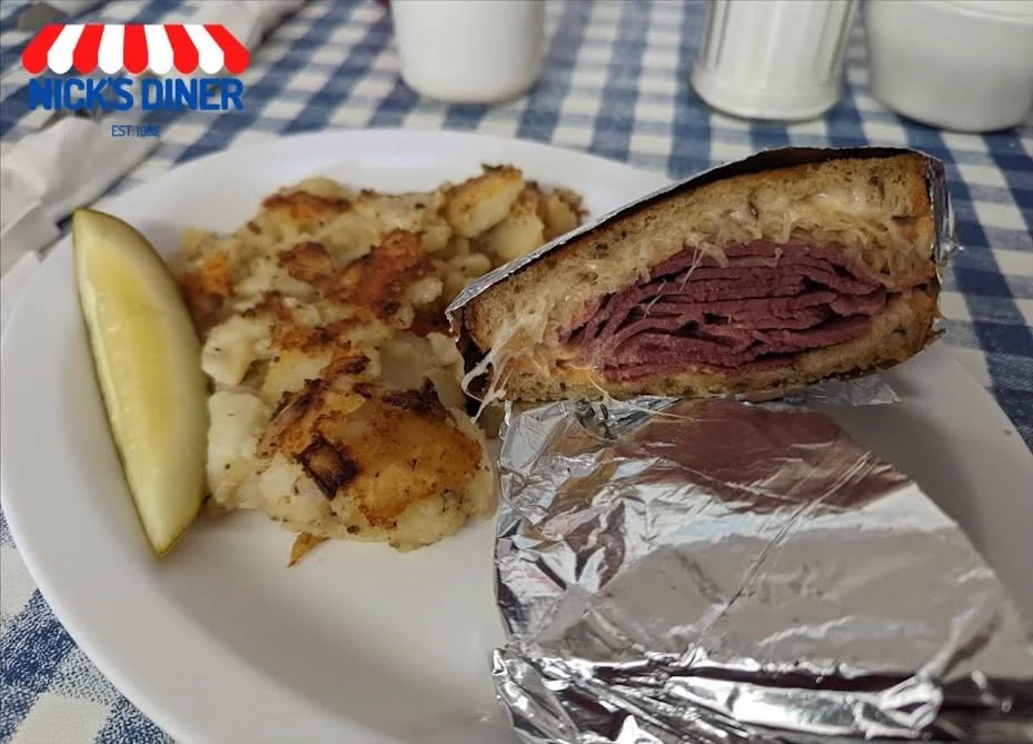 A plate with a corned beef sandwich in foil, a lemon wedge, and a serving of fried fish with a side, set on a checkered blue and white tablecloth.