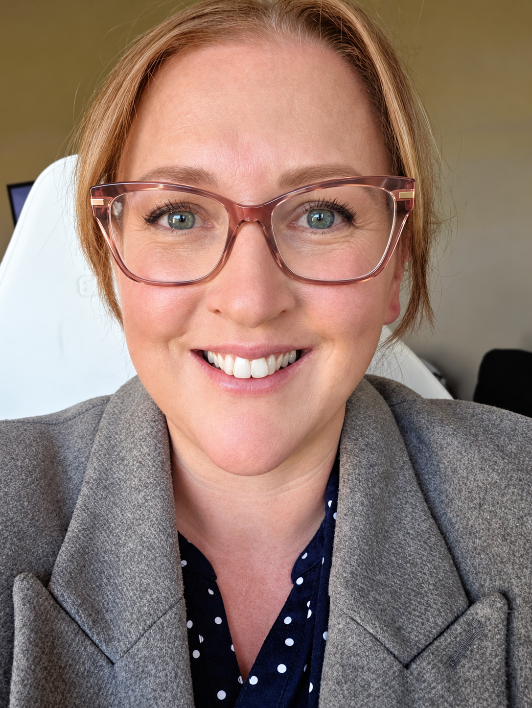 Smiling woman wearing pink eyeglasses, a gray blazer, and a navy polka-dot blouse in a close-up portrait.