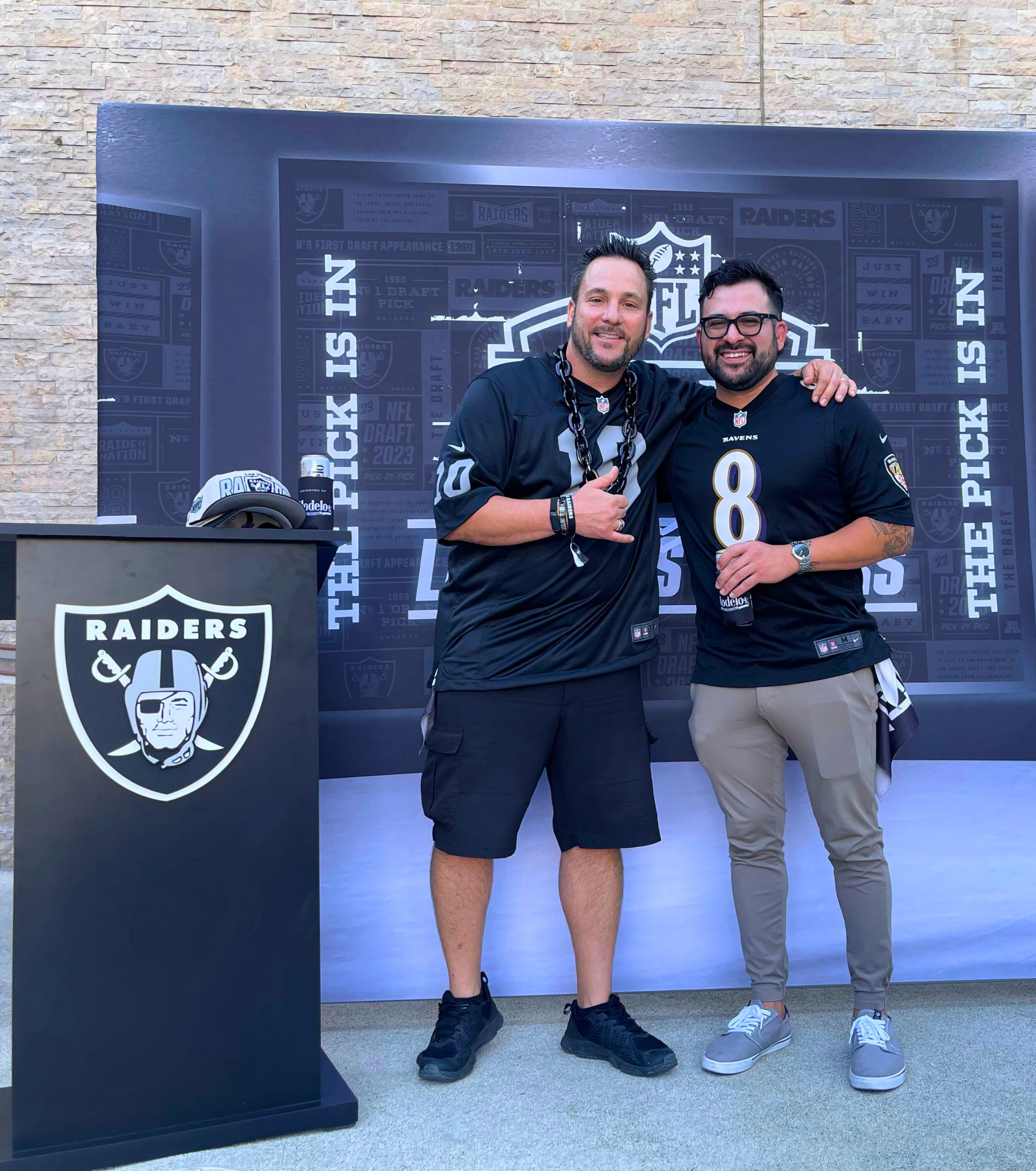 Two men pose in Las Vegas Raiders jerseys beside a large Raiders logo display.