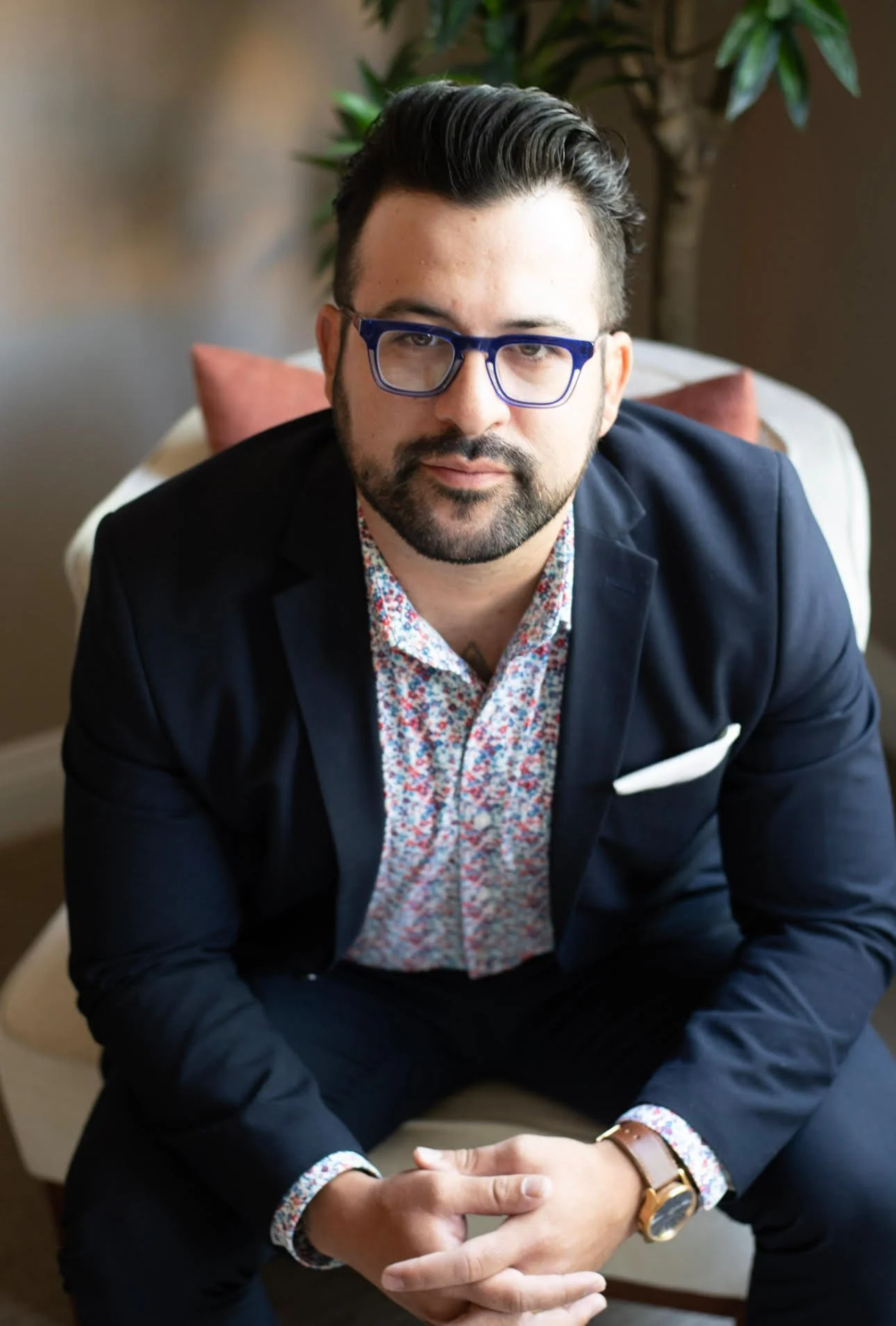 Portrait of a bearded man wearing blue glasses, a navy blazer, and a floral shirt indoors