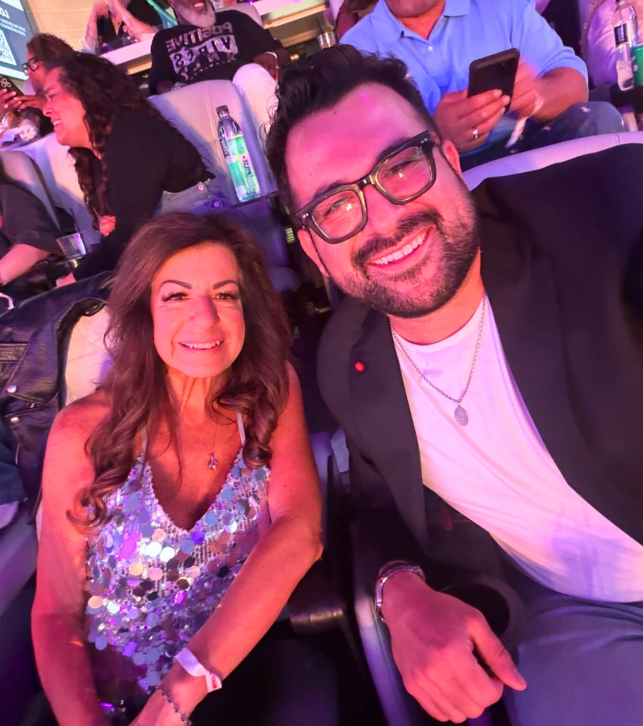 Smiling man in glasses and woman in a sequined dress take a selfie at a brightly lit event surrounded by seated guests.