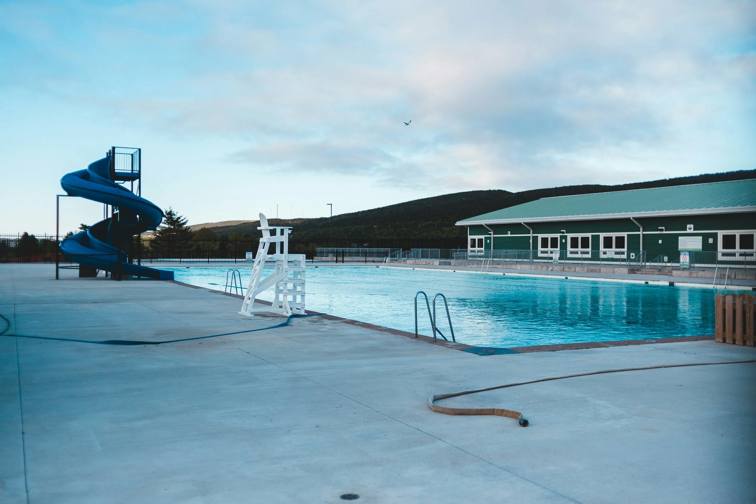 An empty outdoor swimming pool with a blue water slide on the left, a lifeguard chair at the edge, and a building with a green roof in the background, under a partly cloudy sky.