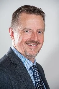 A middle-aged man with short brown hair and a beard, wearing a dark suit, white shirt, and patterned tie, smiling at the camera against a light gray background.