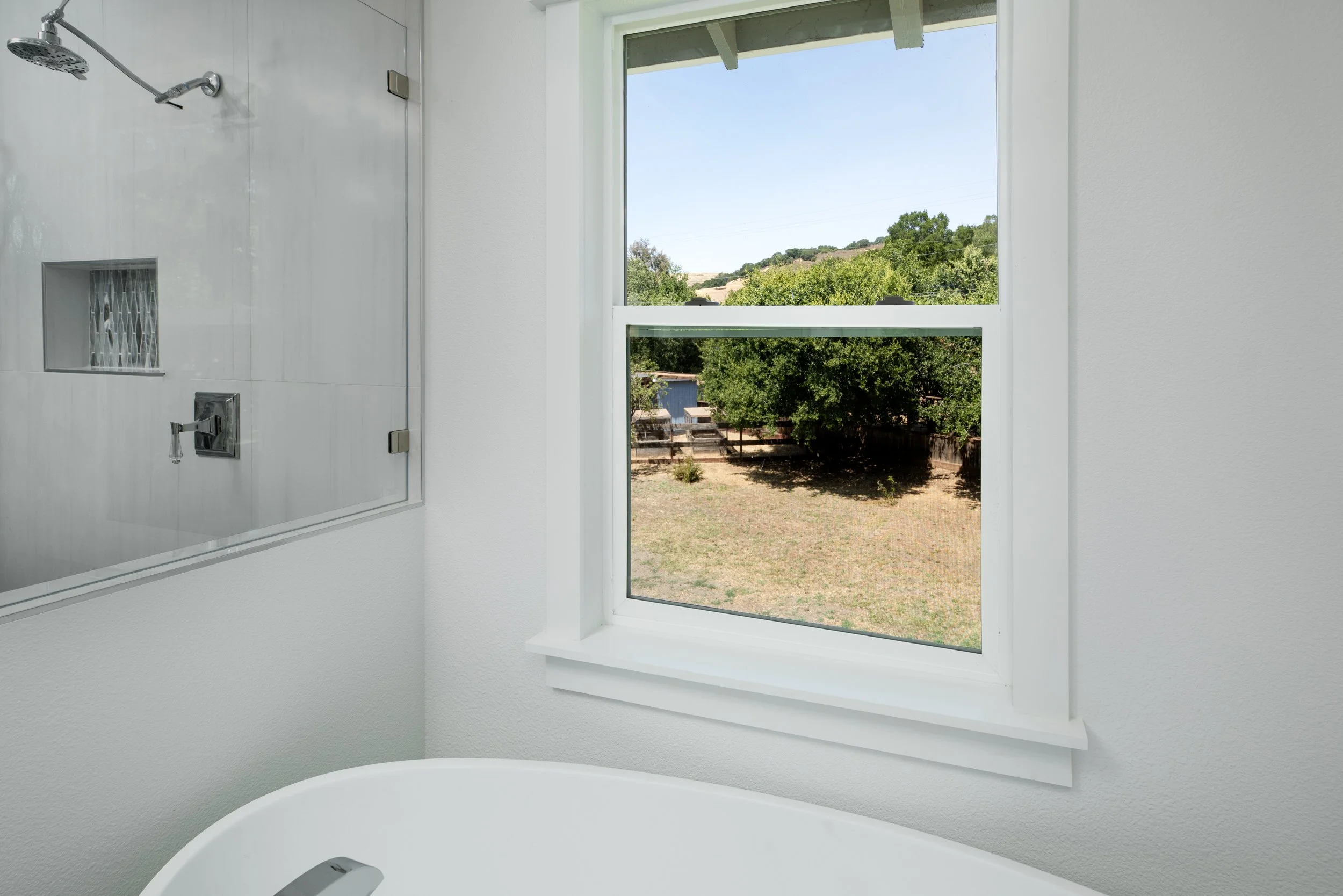 A bathroom with a large window showing an outdoor yard with trees and a wooden fence, a corner of a bathtub, and part of a shower area with white tiled wall and glass enclosure.