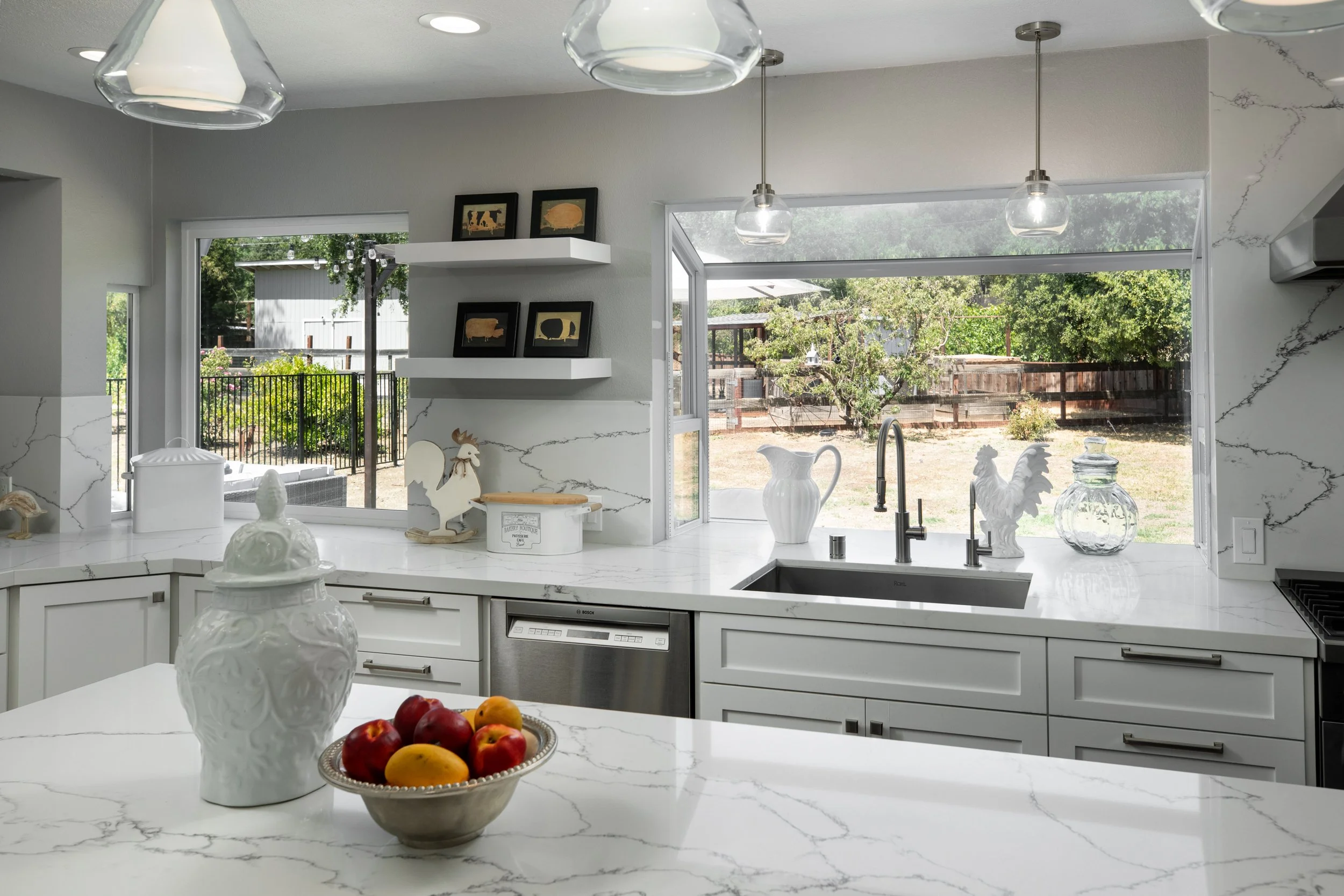 Modern kitchen with a sink, and decorative farm-themed items on the window sill. Large windows show a backyard with a tree and wooden fence.