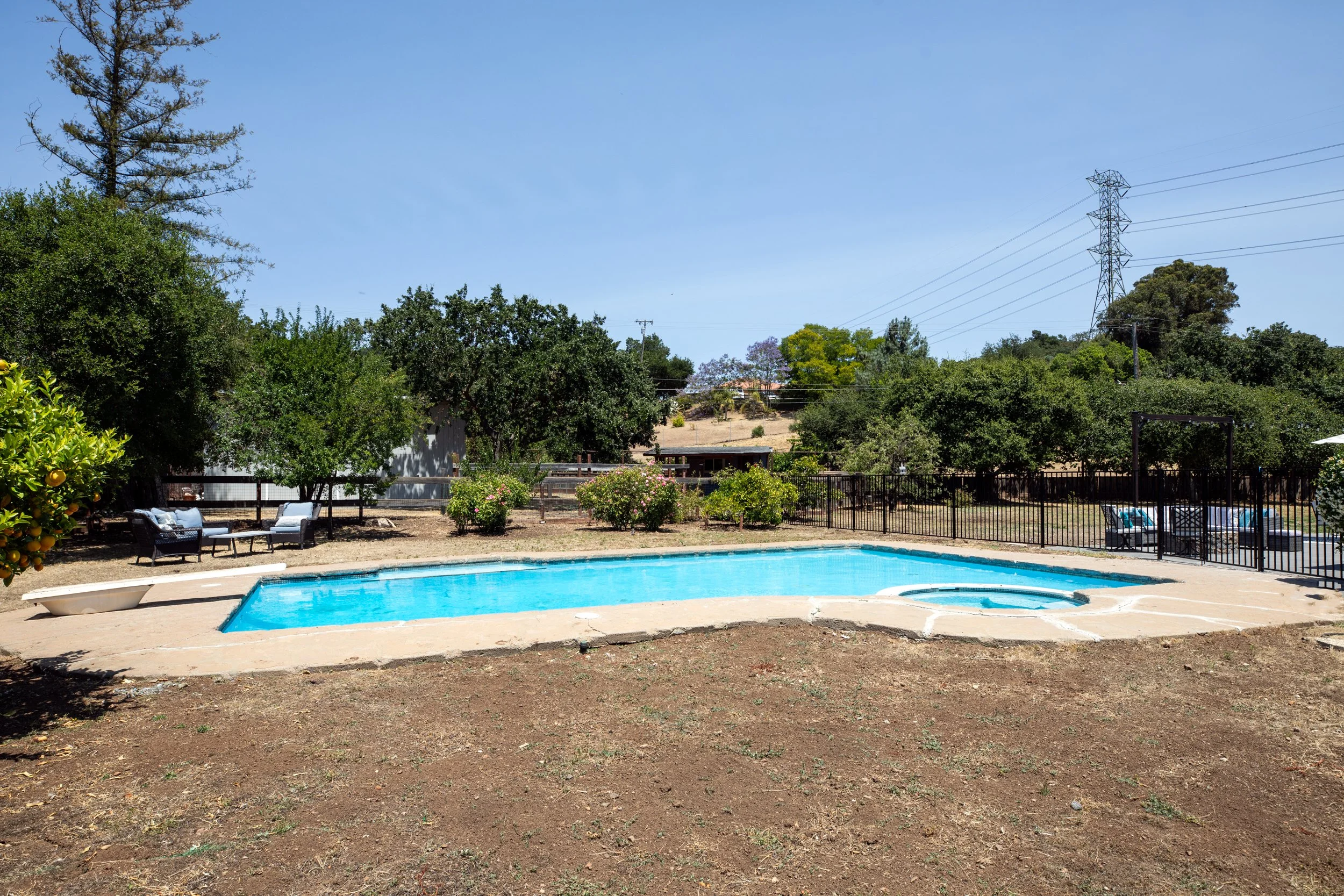 An outdoor swimming pool with a hot tub in a backyard, surrounded by trees, bushes, and outdoor seating on a sunny day.