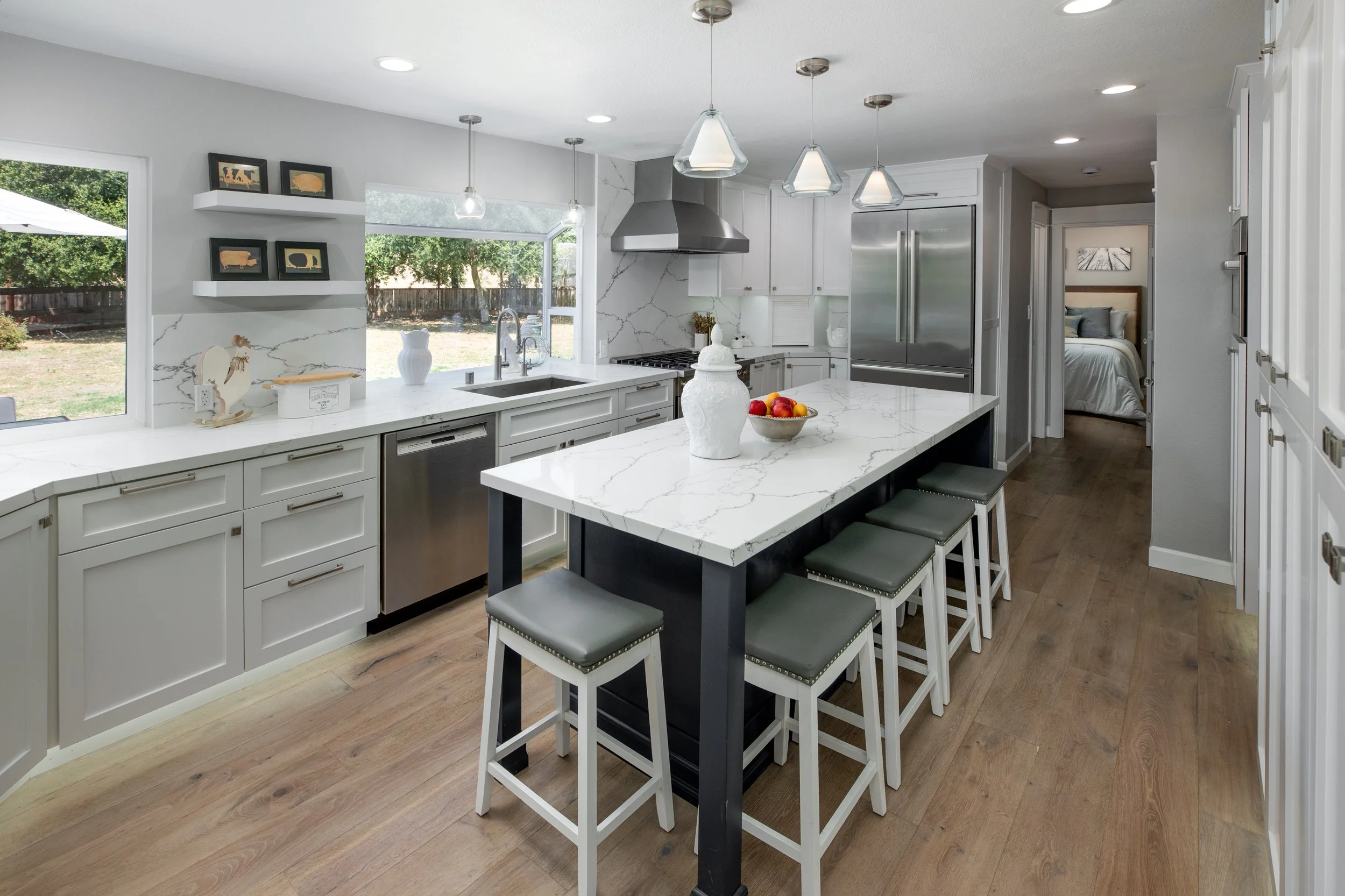Modern kitchen with white cabinets, stainless steel appliances, and an island with seating. Large windows look out onto a backyard.