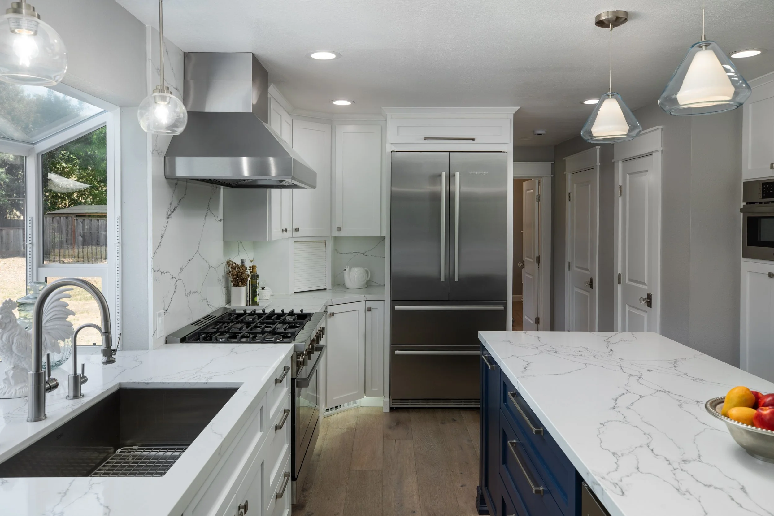 Modern kitchen with white and blue cabinets, a large island with a marble countertop, stainless steel appliances, and pendant lights.