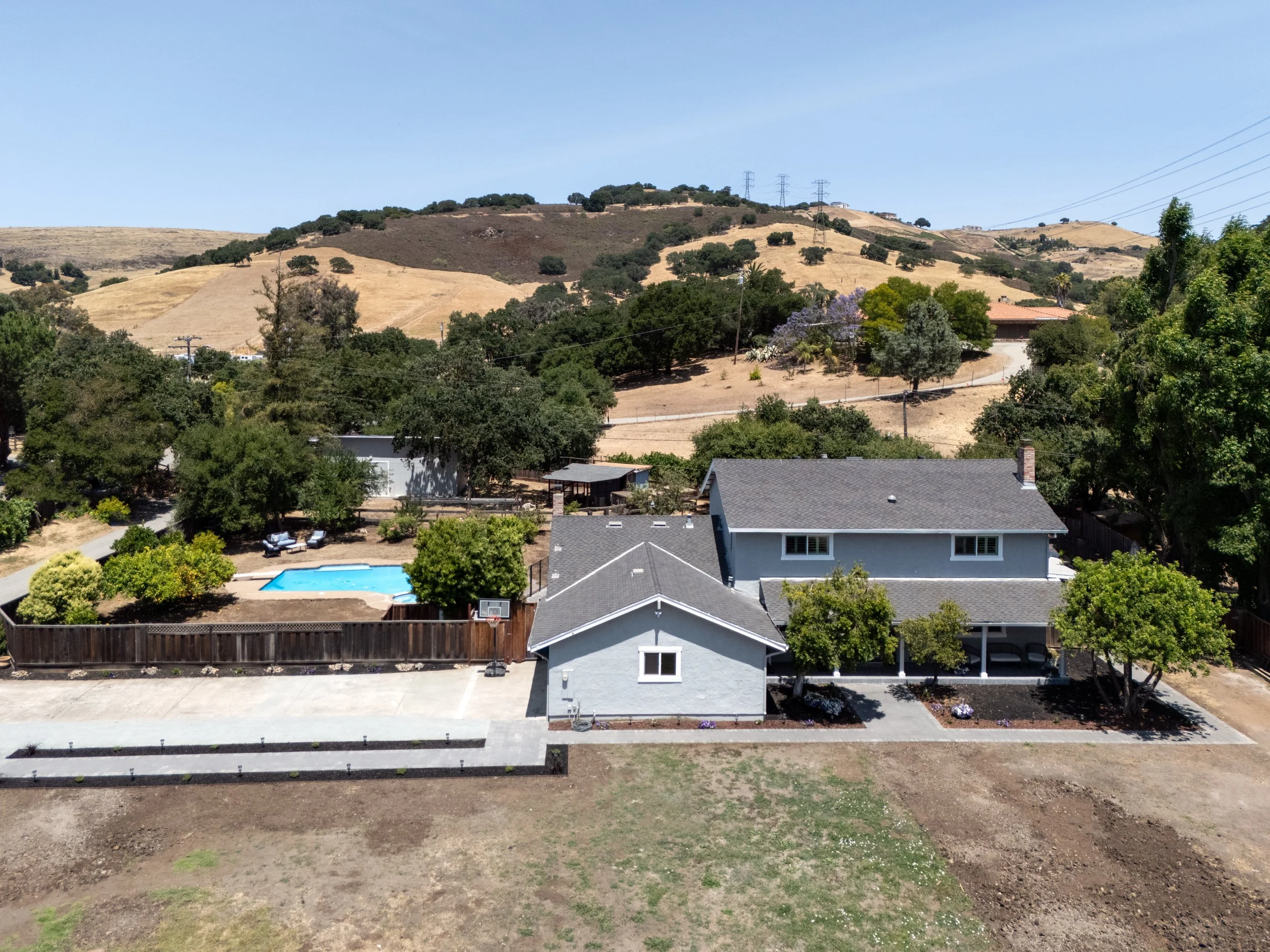 A white two-story house with a gray roof and a covered porch, surrounded by several trees and bushes, with a backyard swimming pool and a basketball hoop, set in a semi-rural landscape with rolling hills in the background.