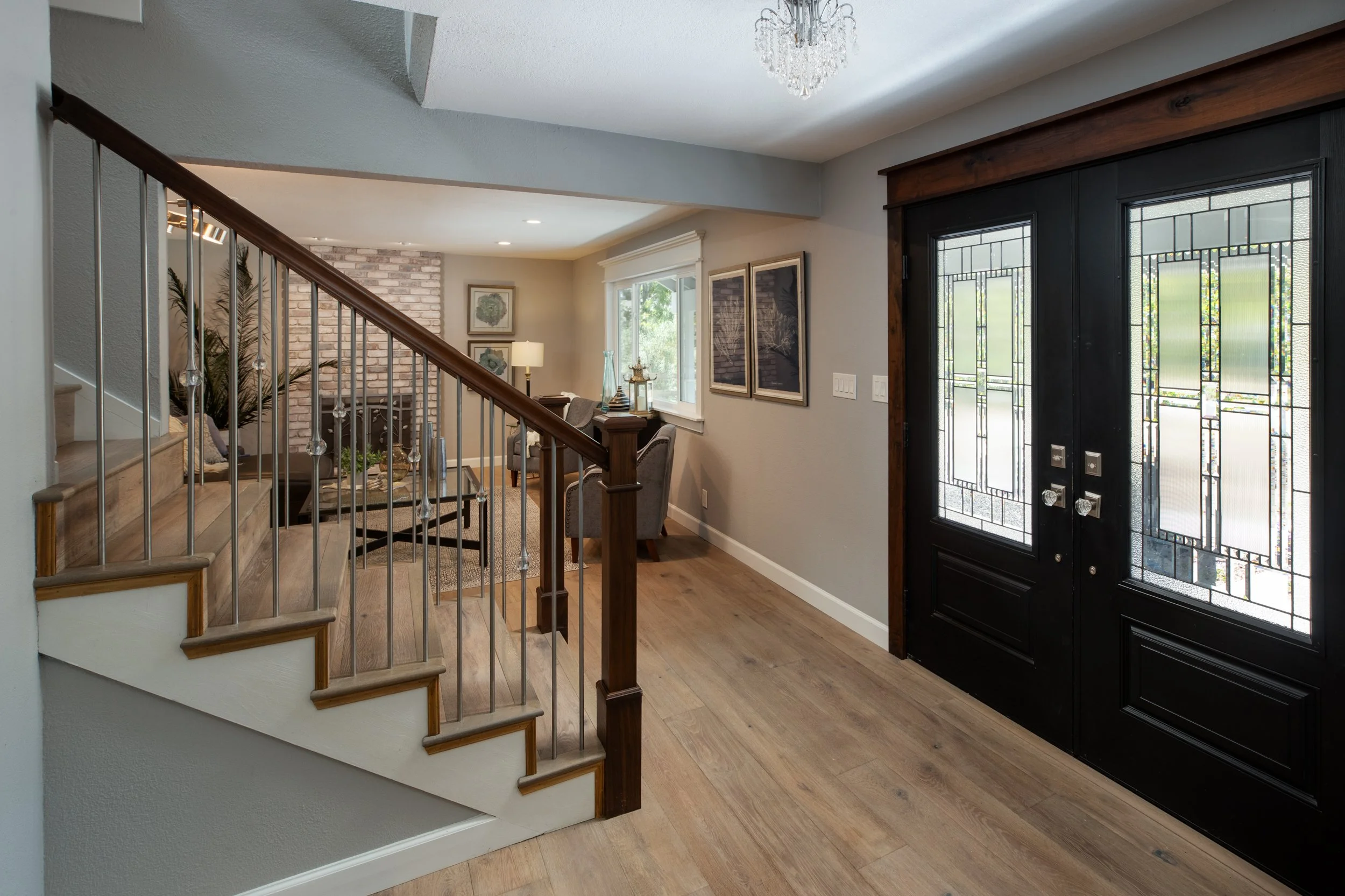Entrance of a house with black double doors with glass panels and decorative design, hardwood flooring, and a staircase with wood steps and metal railing leading to a living room with brick accent wall and furniture.