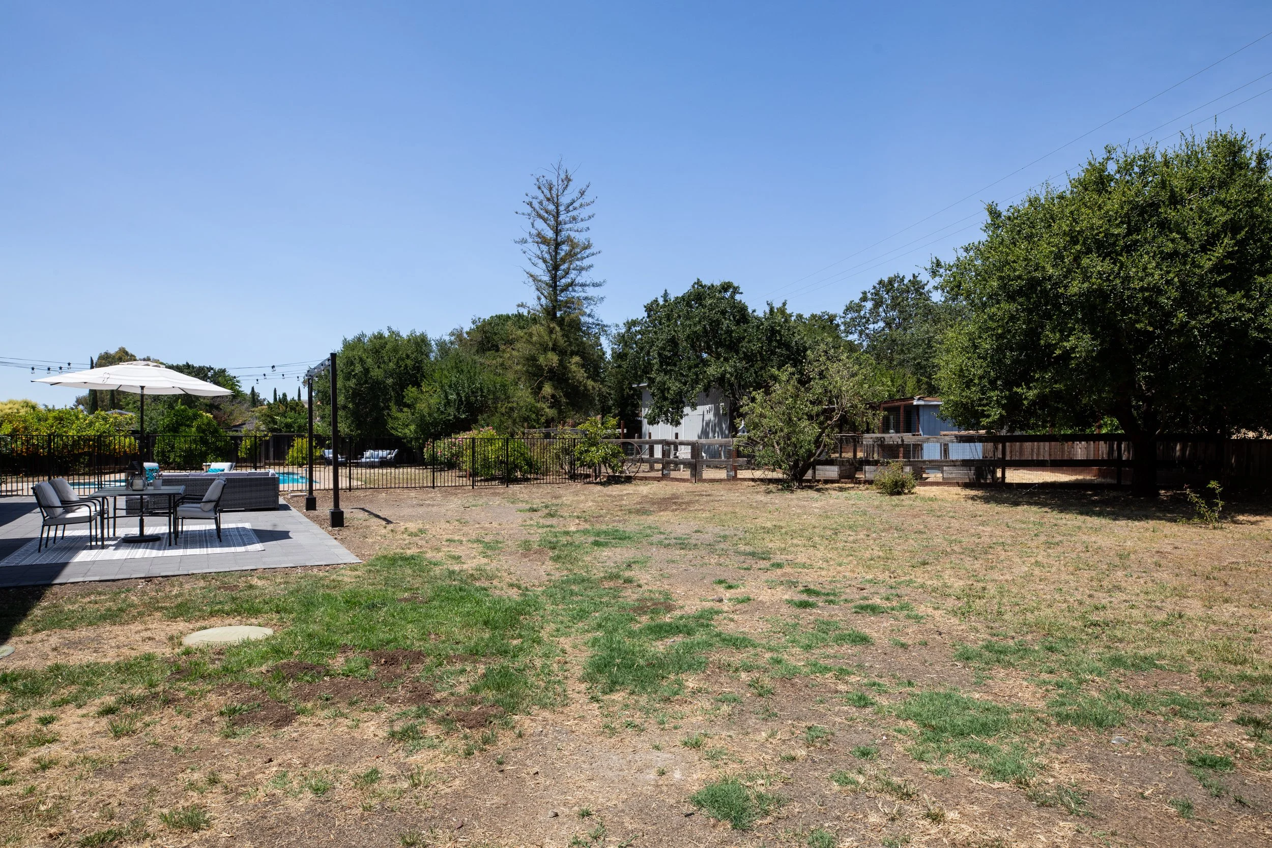 Backyard with a patio area containing an umbrella, table, and chairs, fence, trees, and dry grass on a sunny day.