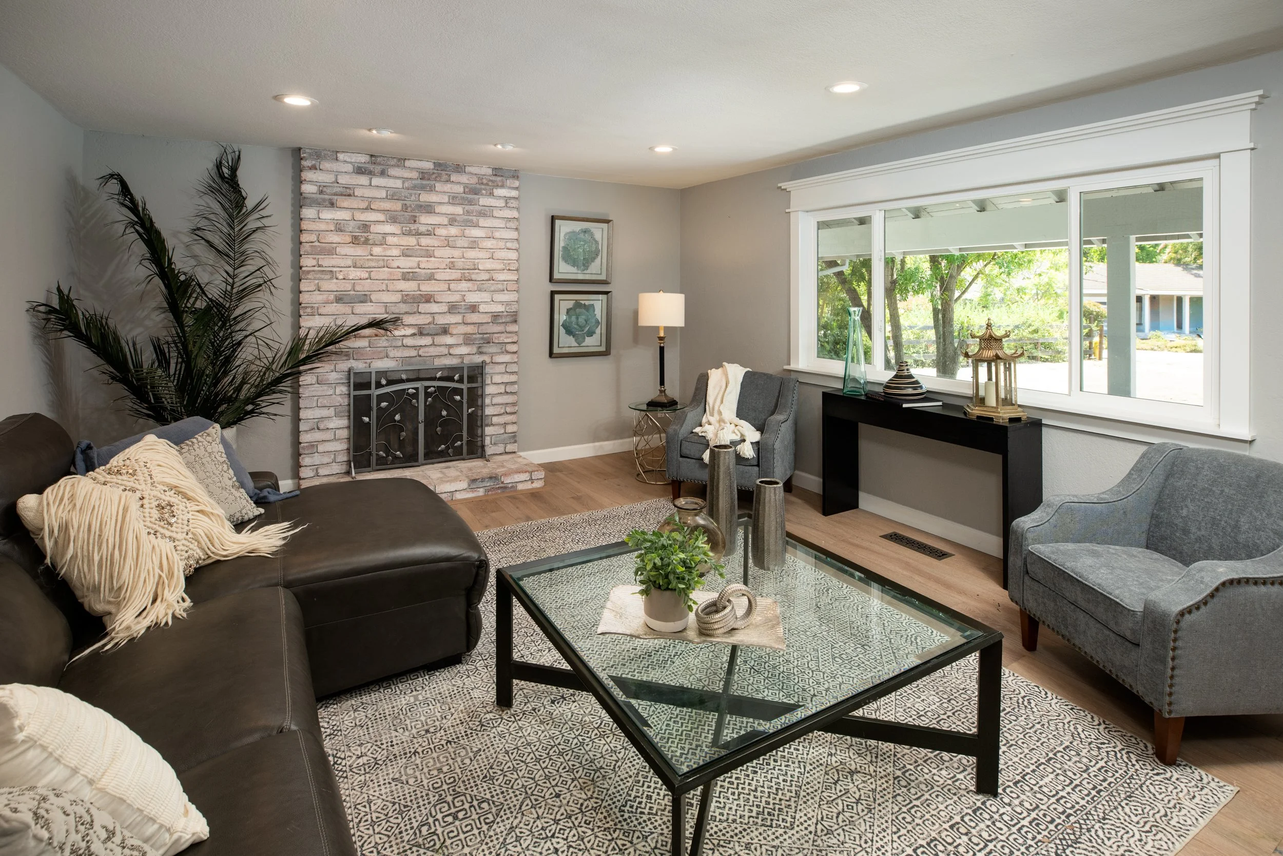 Living room with gray sofa, blue armchair, brick fireplace, large window, and decorative items on black console table
