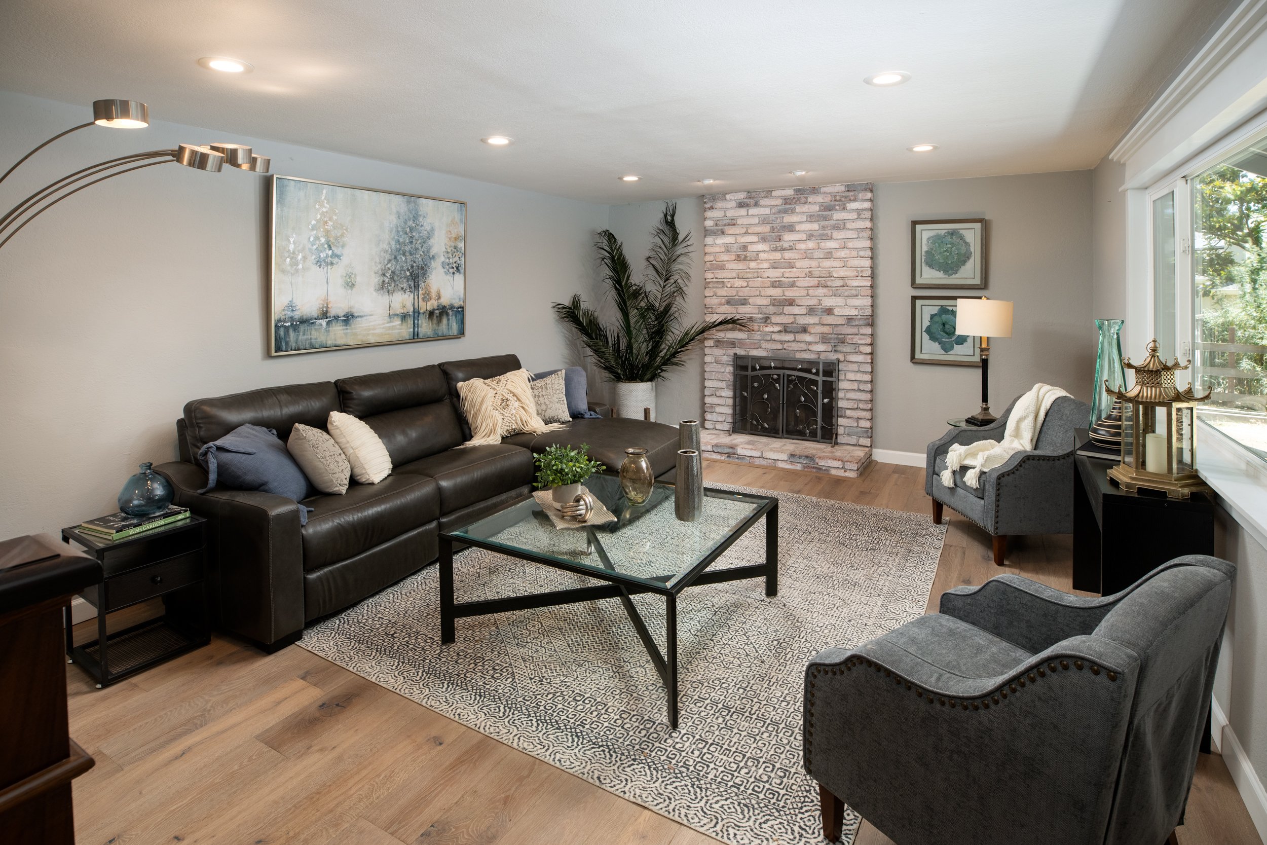 Living room with a brown leather sofa, gray armchairs, a glass coffee table, a brick fireplace, and decor including plants, paintings, and lamps.