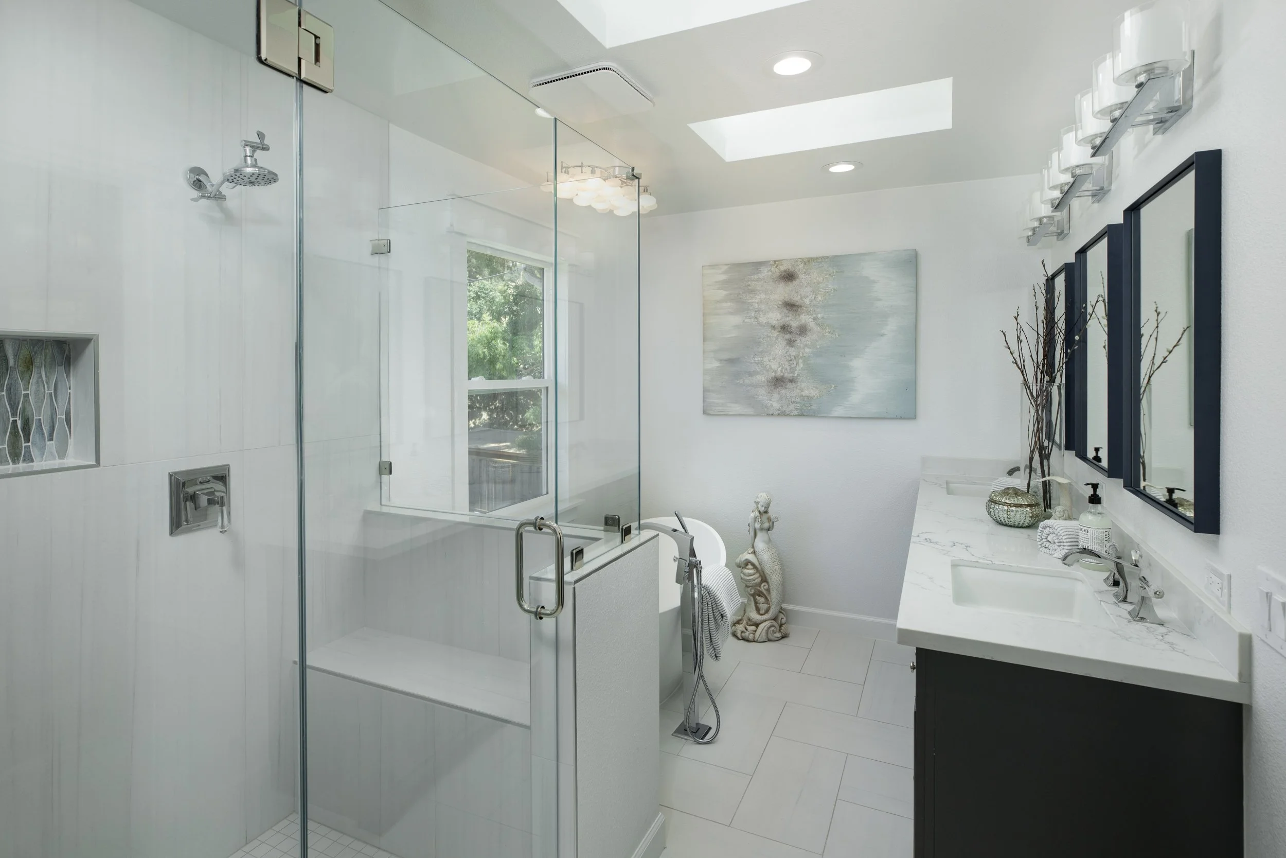Modern bathroom with glass-enclosed shower, white walls, and a double vanity with black cabinets. Decor includes a large abstract painting, vases, and a decorative sculpture near the window. Natural light from a skylight illuminates the space.