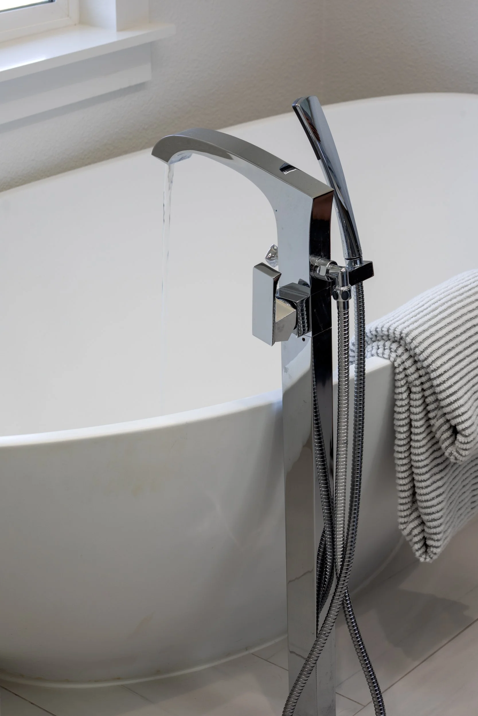 Close-up of a modern chrome bathtub faucet with a water stream, attached to a white bathtub with a folded gray and white striped towel over the edge.