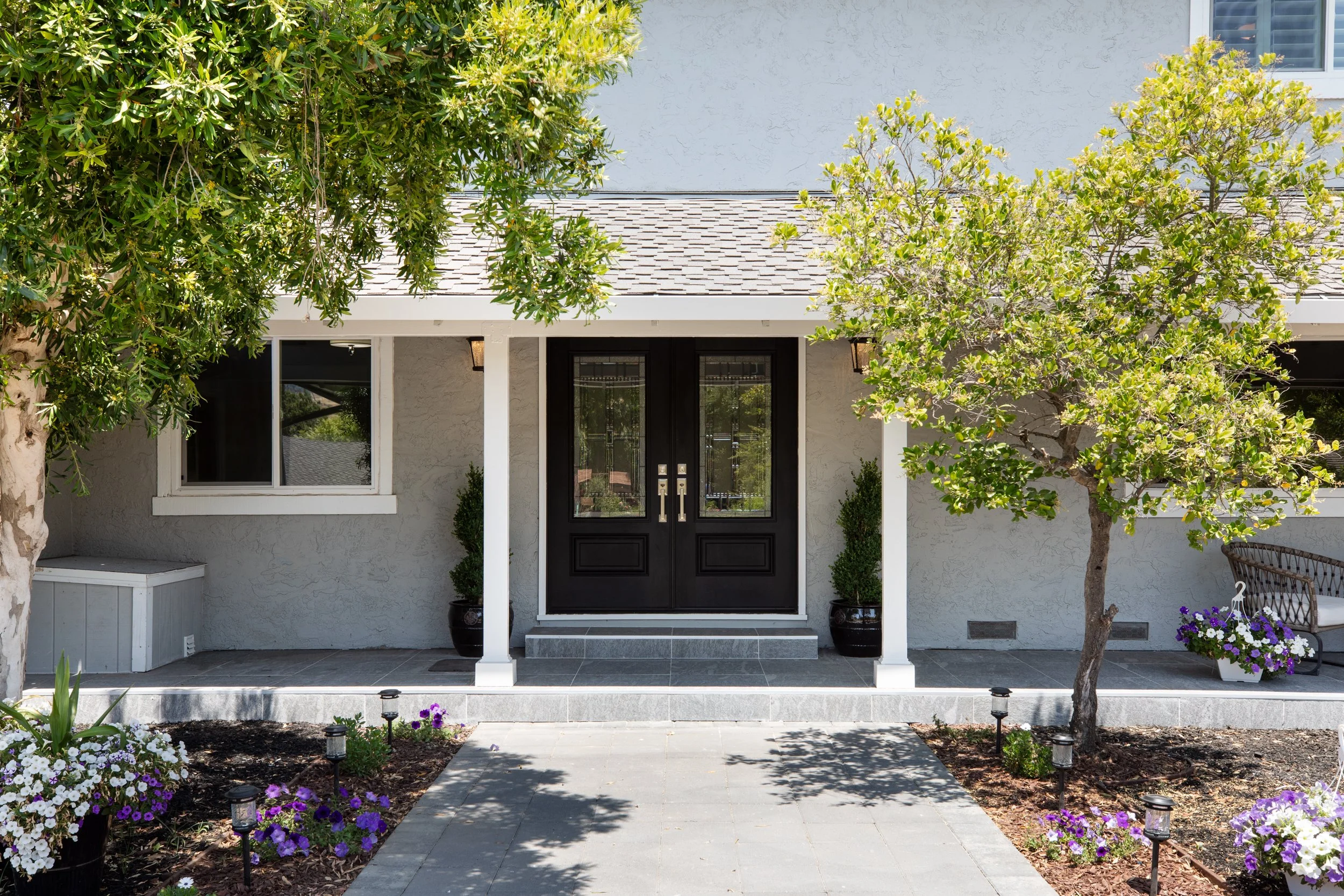 Front entrance of a house with a black double door, two potted plants, and a porch with gray tiles. Two trees and flower beds with purple and white flowers in pots and planted in the soil decorate the front yard. Small pathway stones lead to the porc