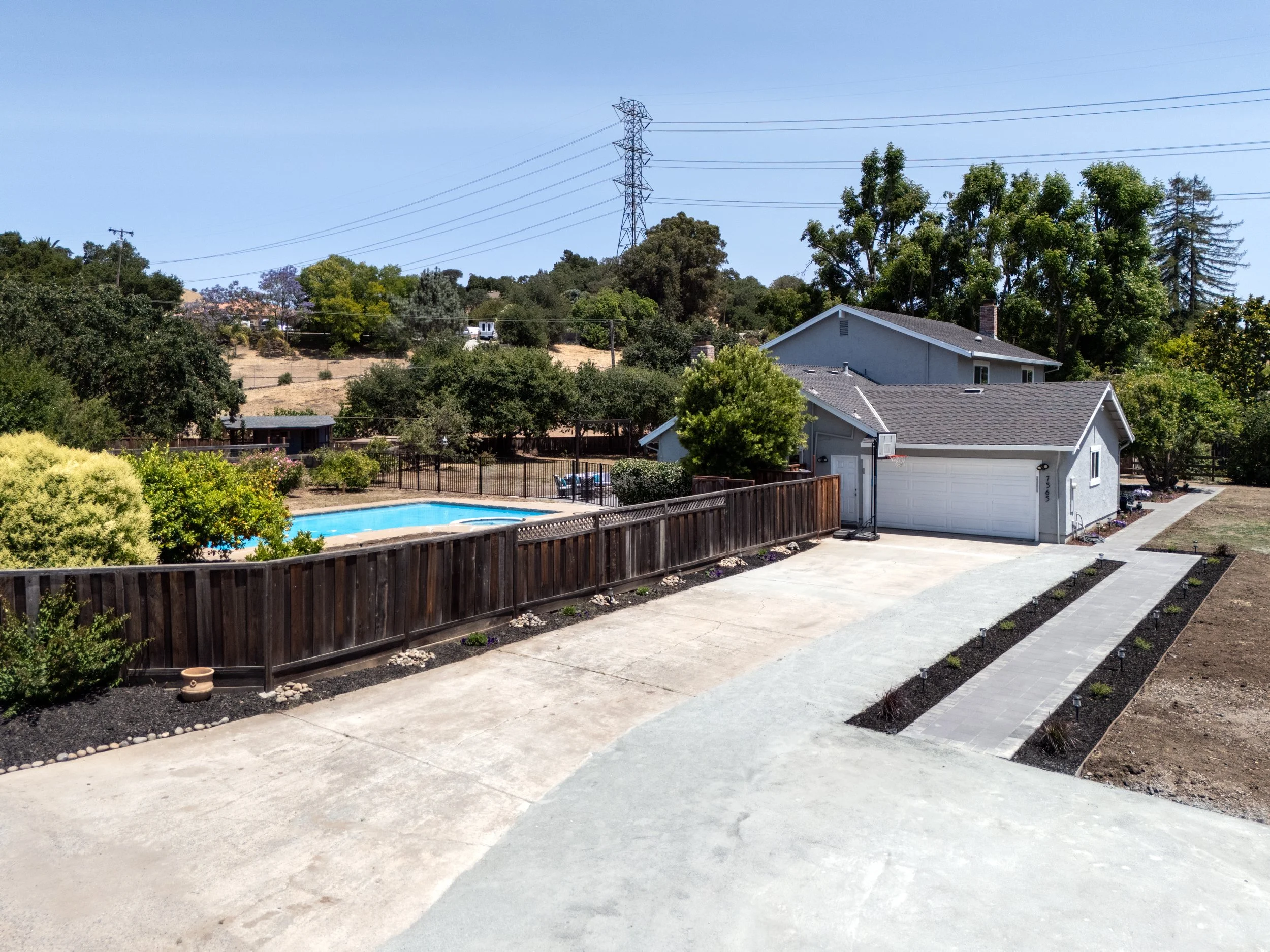 A backyard with a concrete driveway, a blue swimming pool, a gray house with a garage, a basketball hoop, and landscaped garden beds.