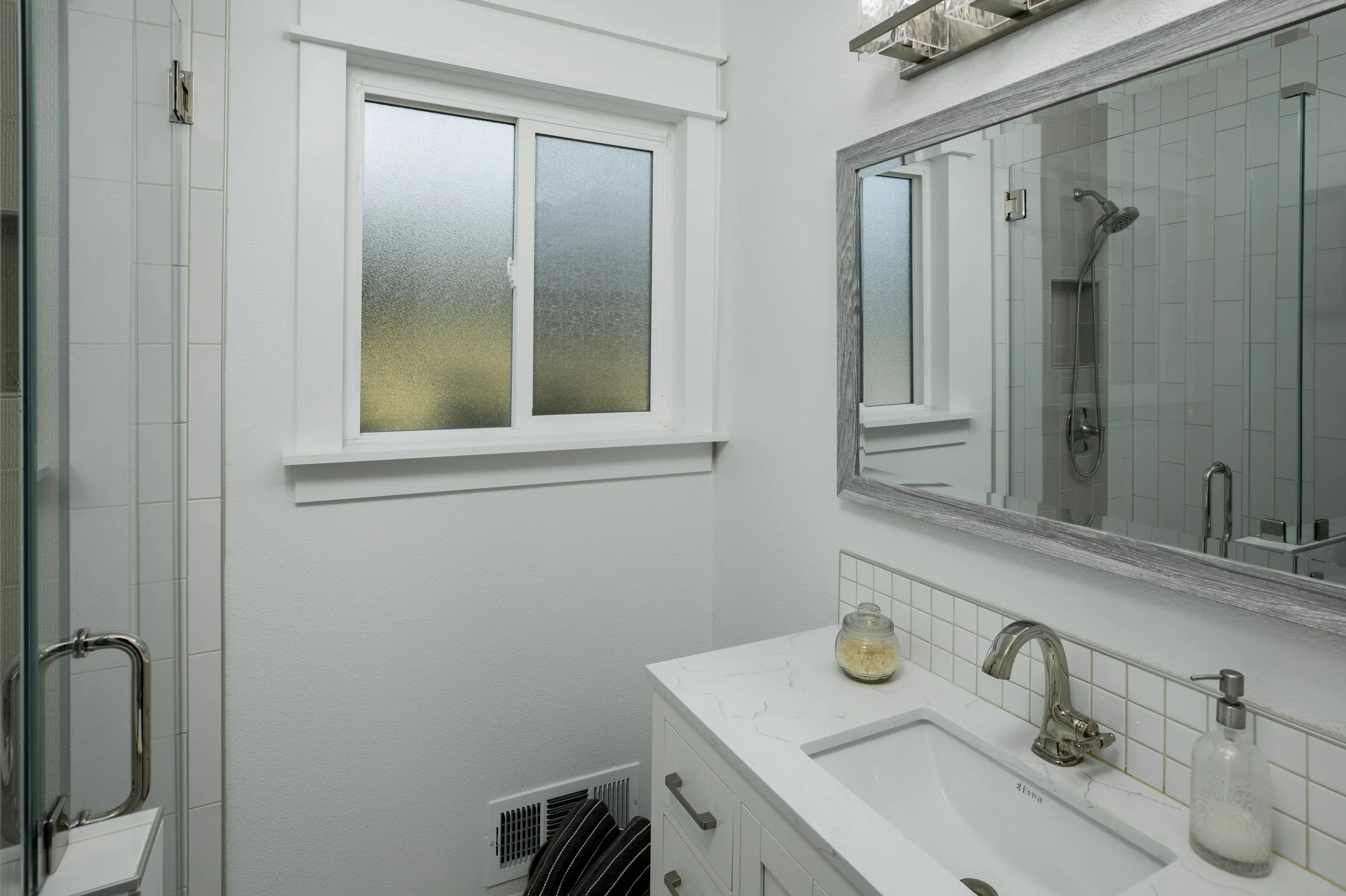 A small bathroom with a white vanity, large mirror, frosted window, and a glass-enclosed shower with a showerhead.