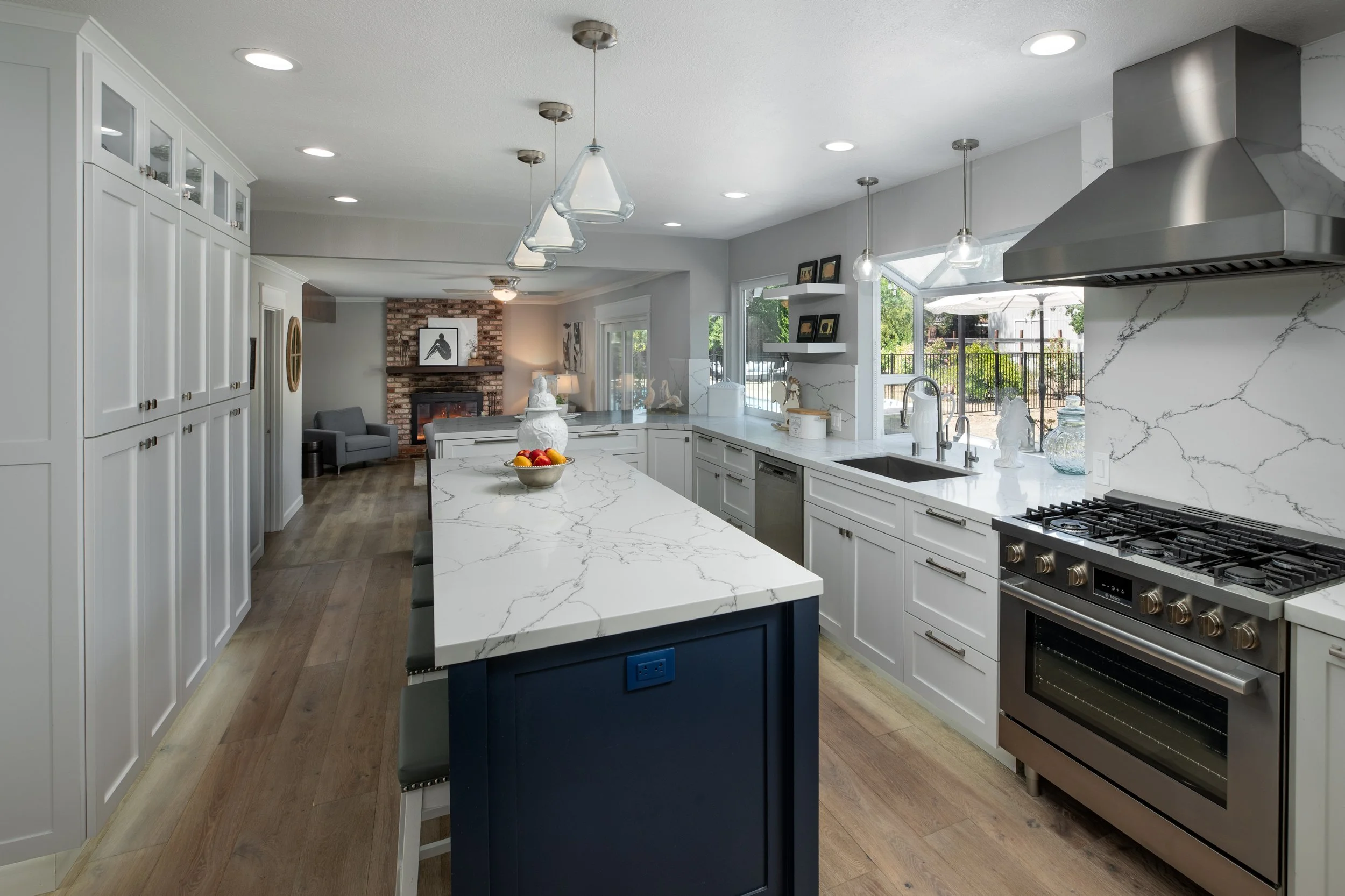 Modern kitchen with white cabinetry, stainless steel appliances, and a central island with a blue base and bar stools. There is a brick fireplace and living area visible in the background.