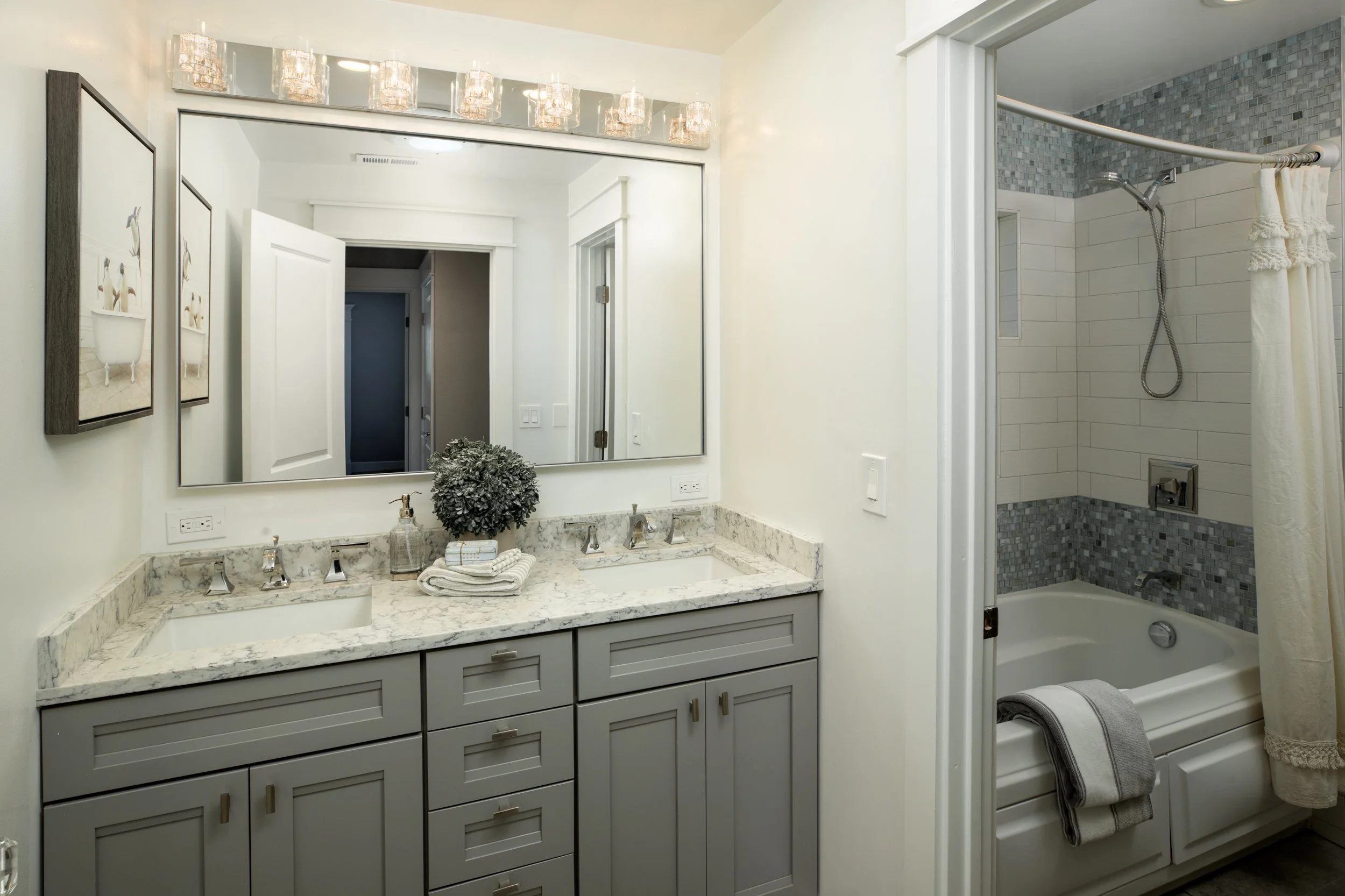 A modern bathroom with a double vanity featuring two sinks, and a large mirror. To the right, a bathtub with a shower and a curtain, surrounded by small gray tiles.
