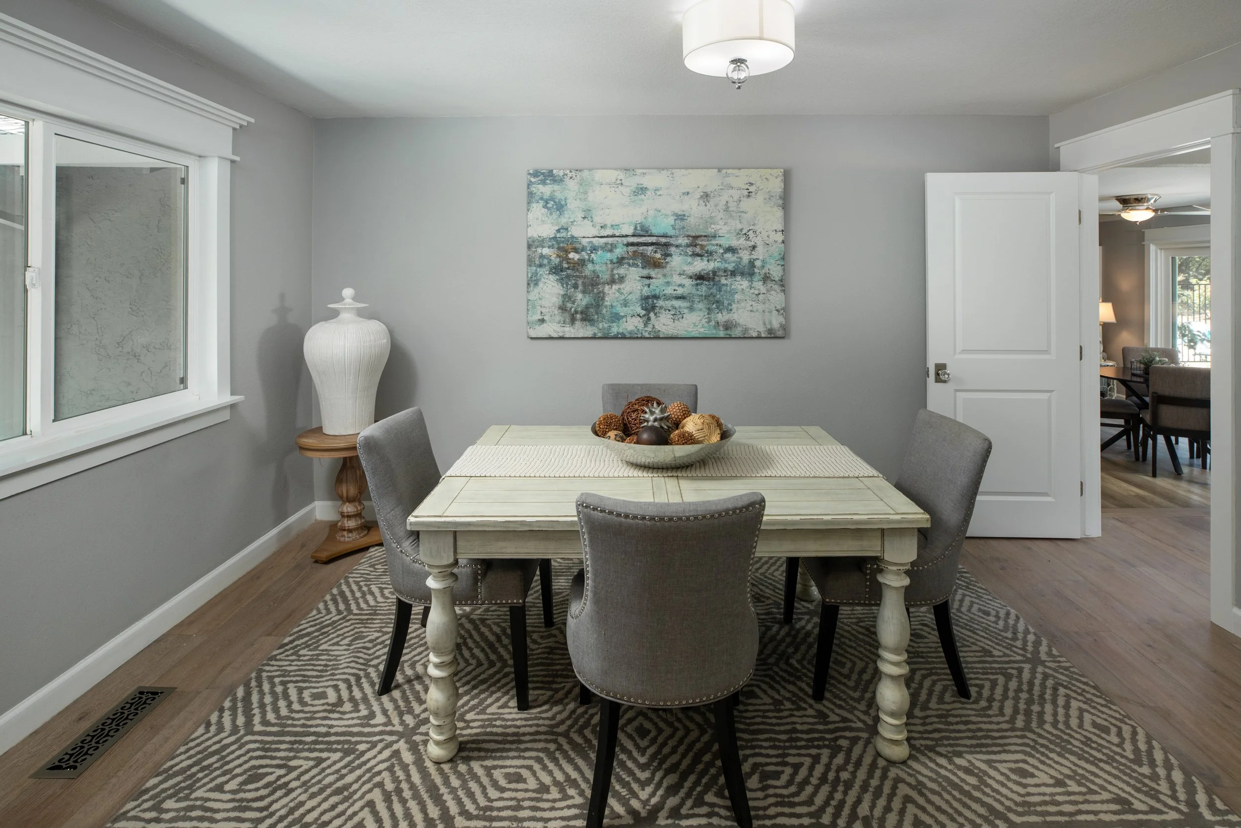 Dining room with a rectangular table, six gray upholstered chairs, a large white decorative vase on a wooden side table, an abstract painting on the gray wall, a patterned rug, and a bowl of decorative spheres.