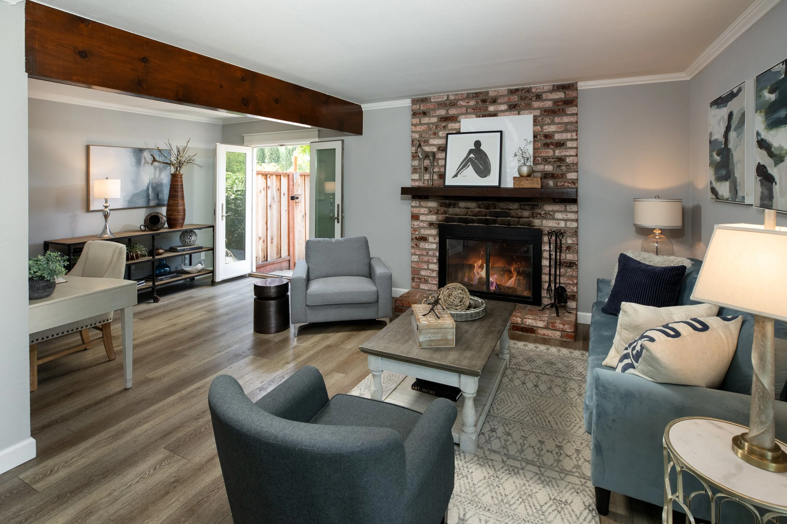 Cozy living room with a brick fireplace, gray and blue furniture, and a view of the open door leading outside.