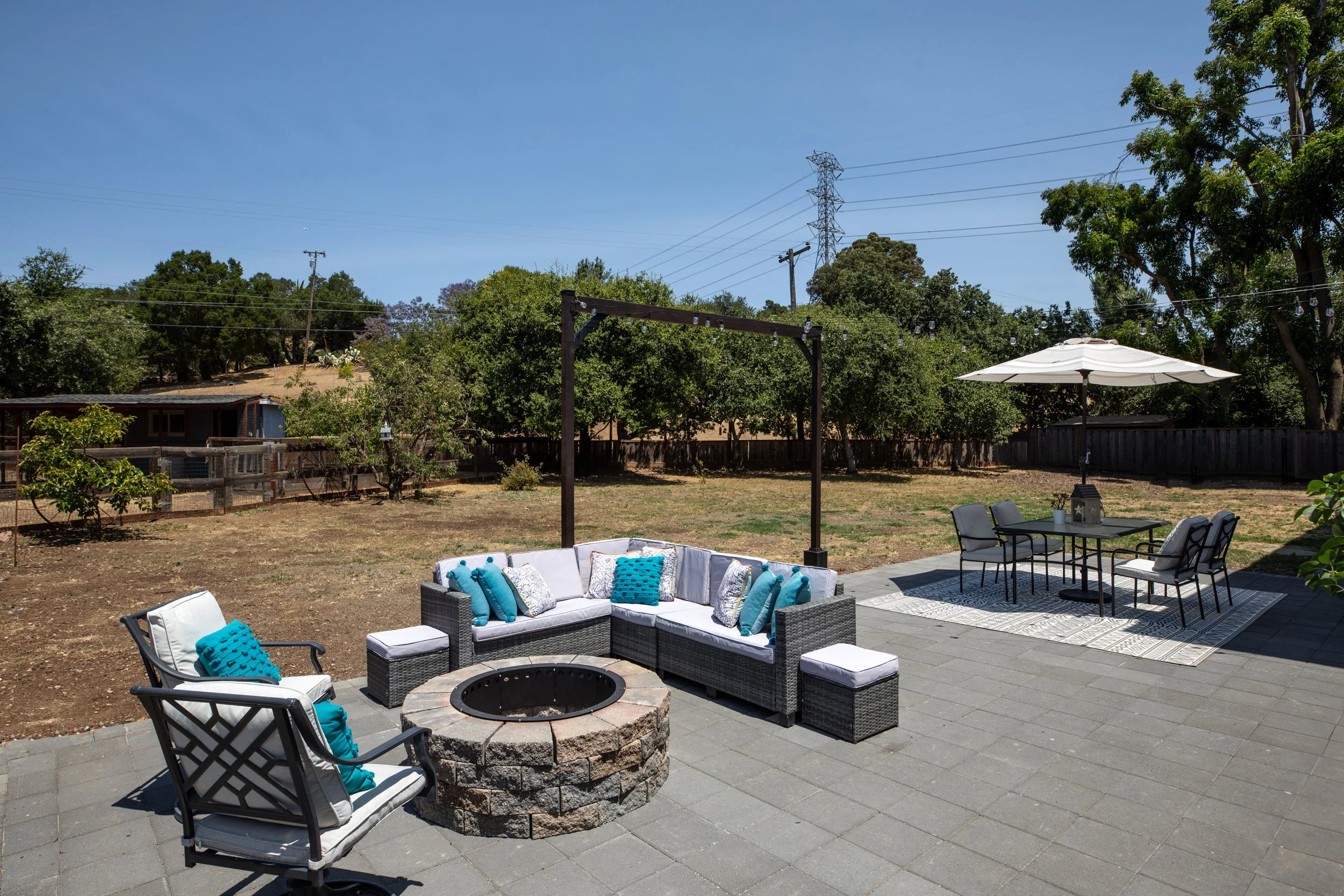 Backyard patio with outdoor furniture, including a firepit, sectional sofa with blue pillows, and a dining table with chairs and an umbrella, on a sunny day with trees and power lines in the background.