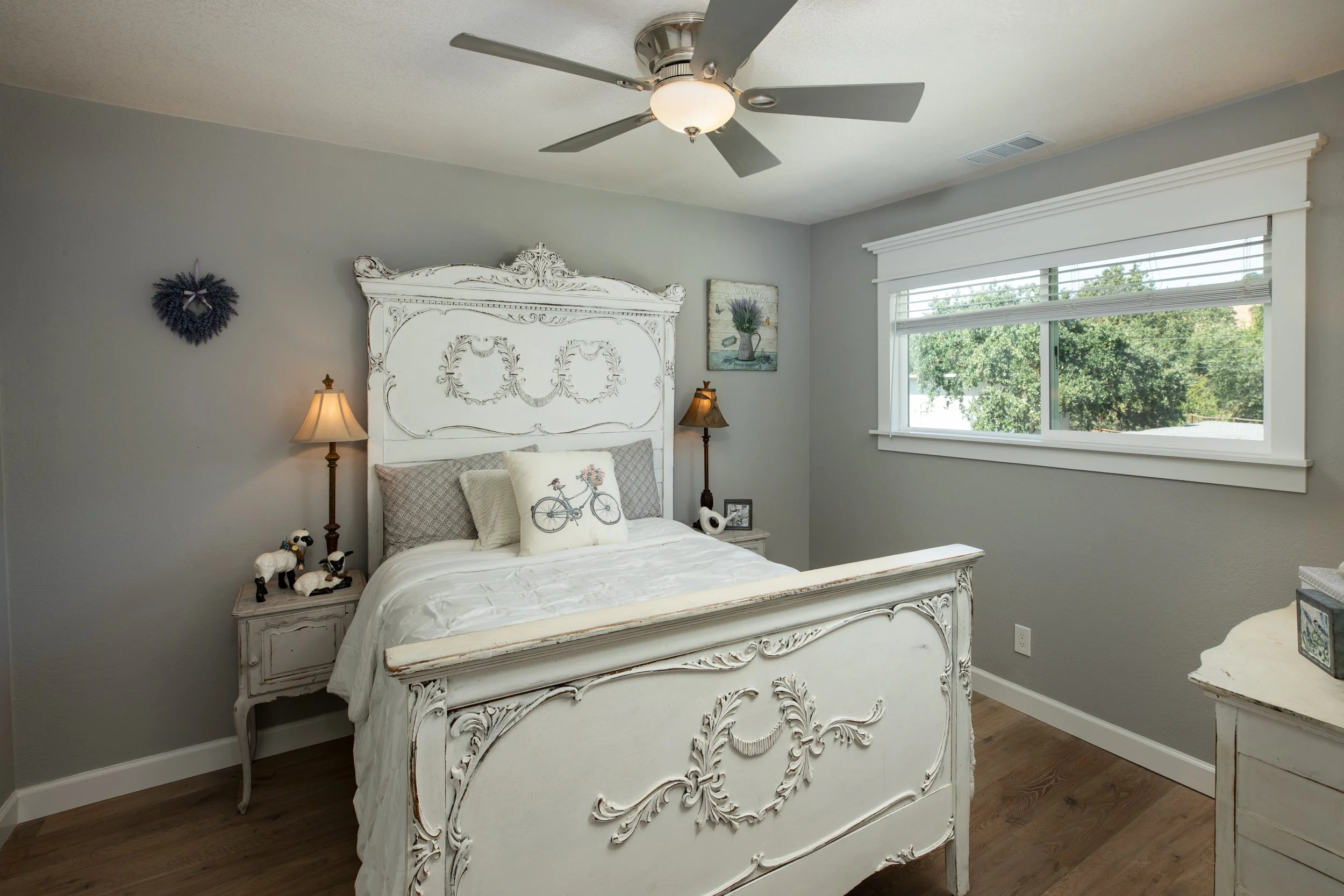 A bedroom with a white ornate bed, gray walls, large window with white trim, ceiling fan, bedside lamps, decorative pillows, and wall art of flowers.