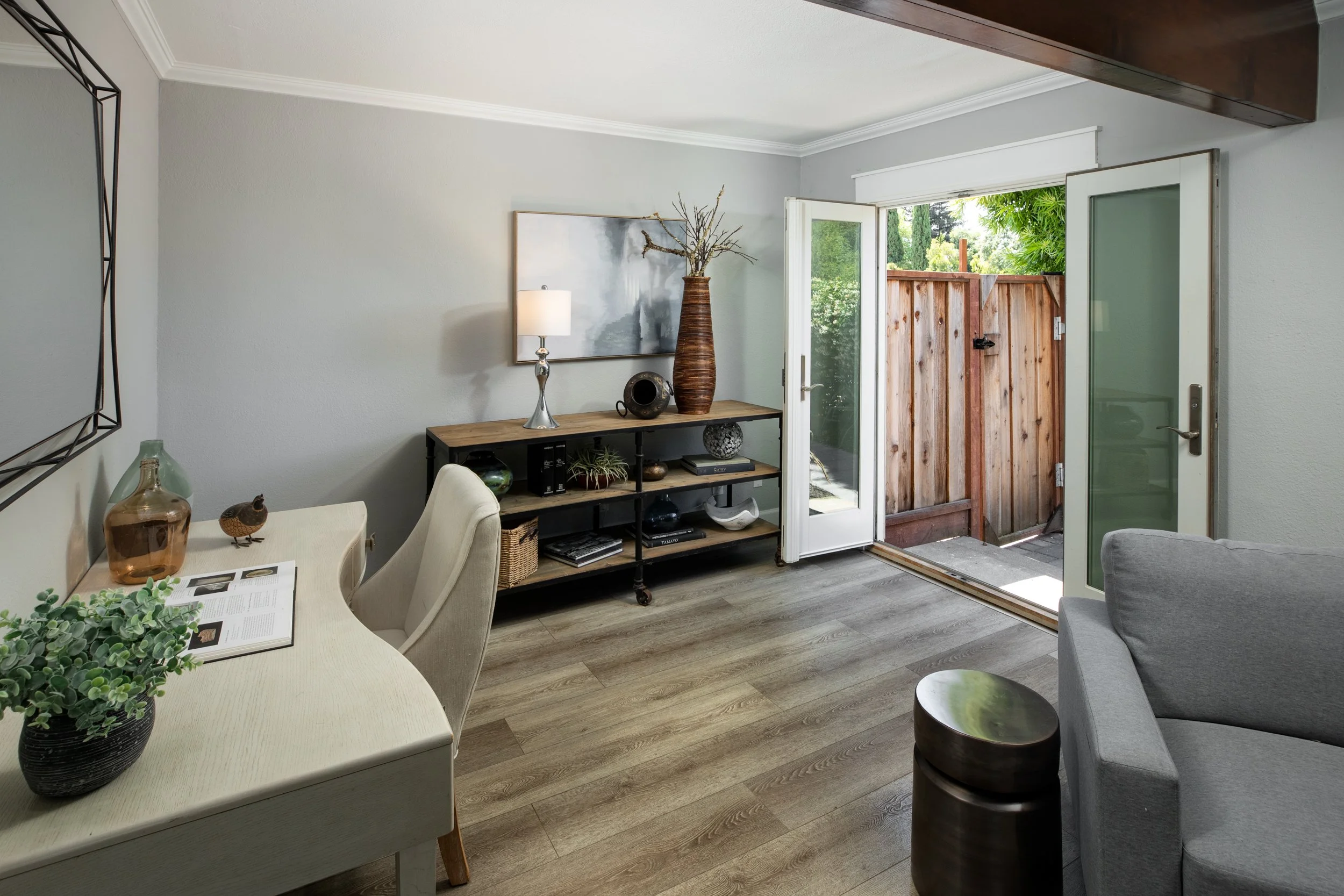 Living room with open door leading outside, decor includes a bookshelf, a lamp, vases, and a plant, with wood flooring and light gray walls.