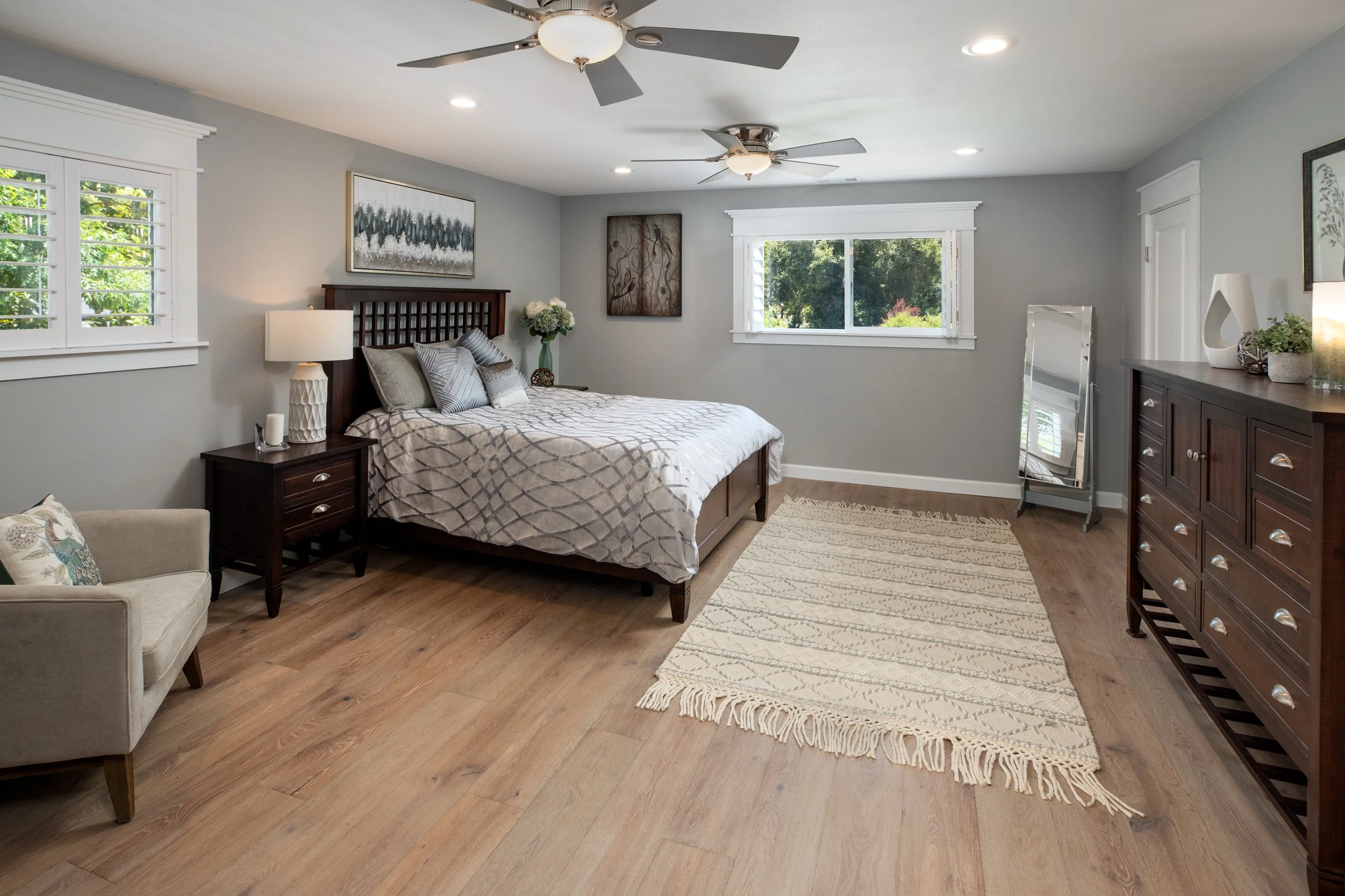 A spacious bedroom with a bed, nightstands, a dresser, a small armchair, and windows with white shutters. The room has hardwood floors, ceiling fans, and decor including paintings and vases.